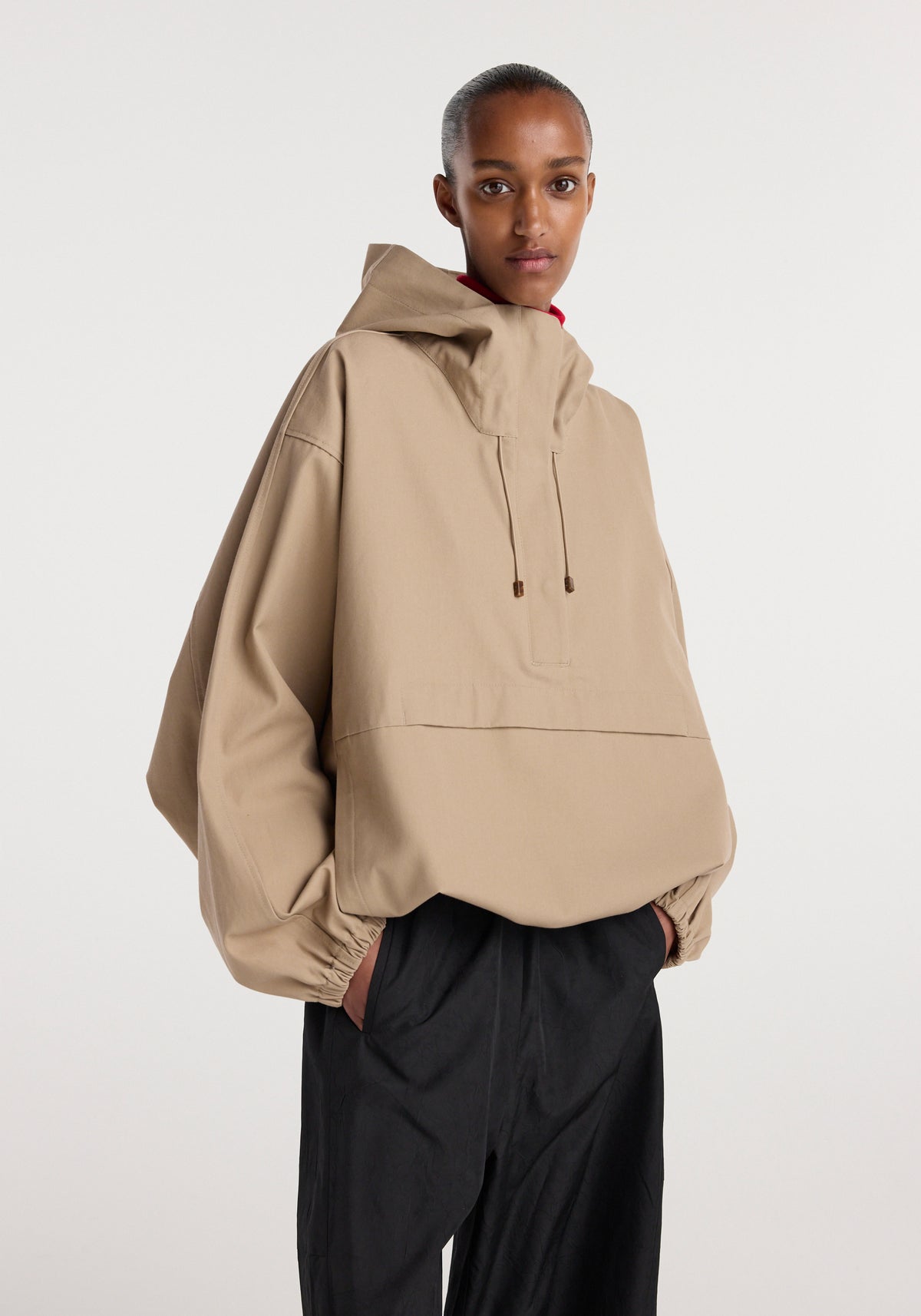 A model wearing a beige oversized hooded anorak with drawstrings and elastic cuffs paired with black pants, standing against a plain white background.
