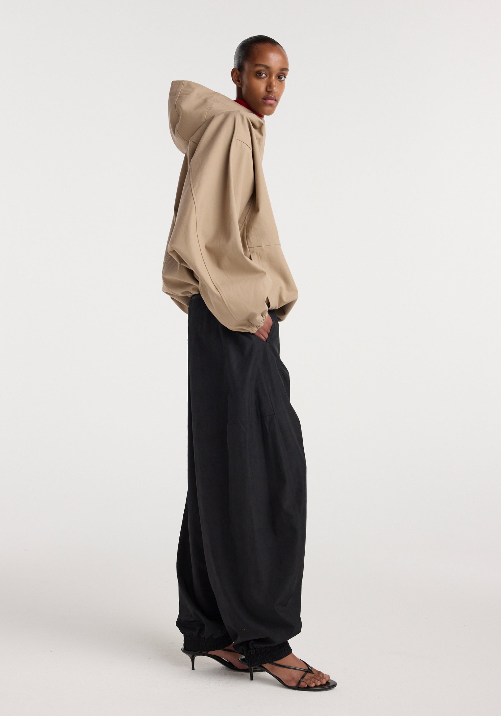 Side view of a model wearing a beige hooded anorak with oversized sleeves paired with loose black wide-leg pants and black strappy heeled sandals.