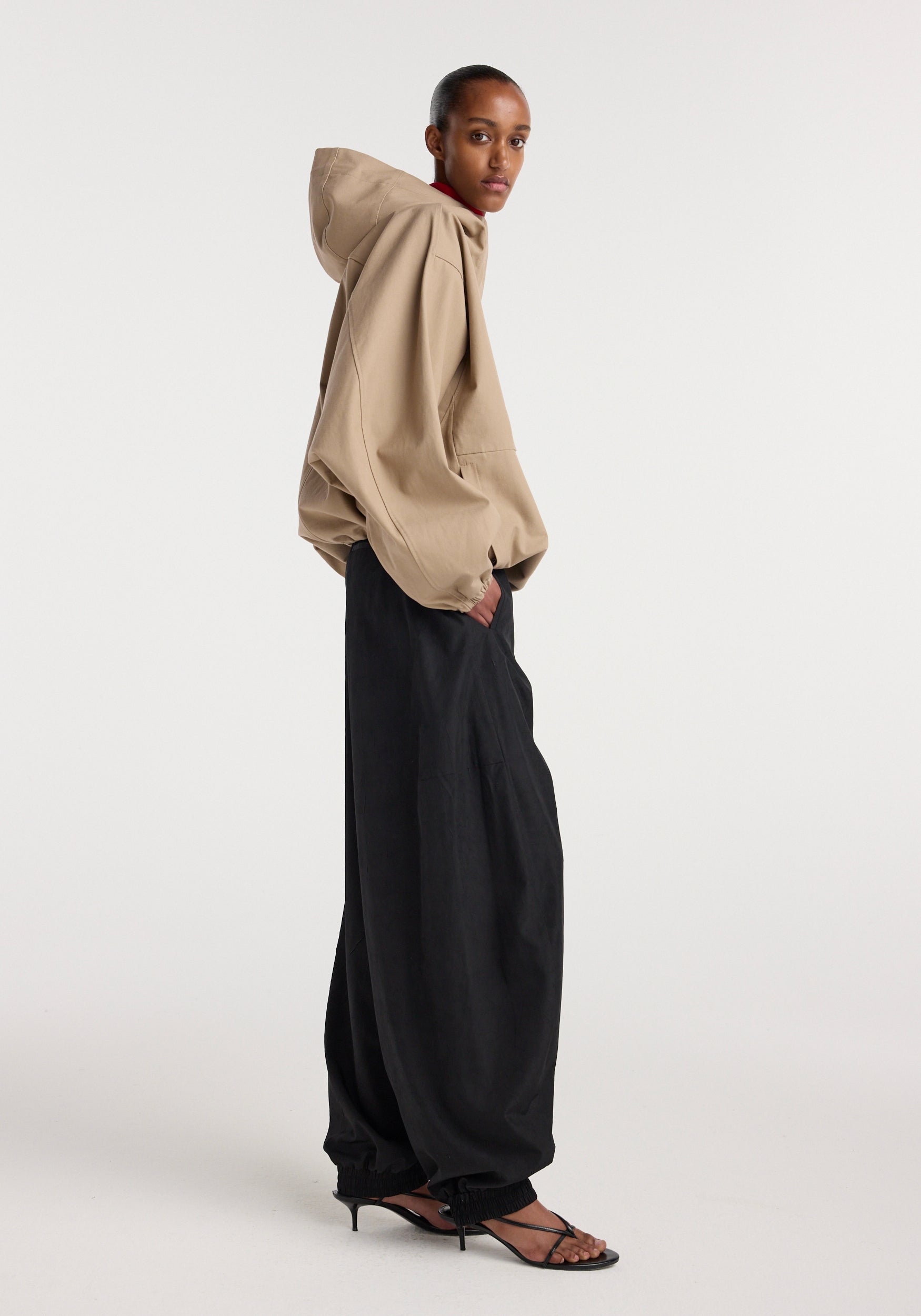Side view of a model wearing a beige hooded anorak with oversized sleeves paired with loose black wide-leg pants and black strappy heeled sandals.