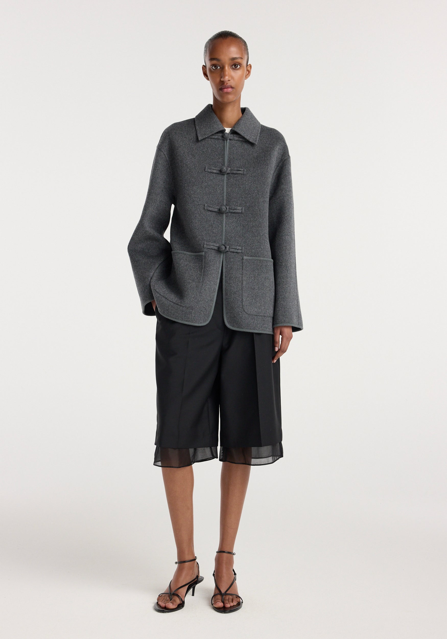 Model wearing a dark grey melange double-faced wool pankou closure jacket with large front pockets and wide sleeves, paired with black knee-length shorts and strappy heels.