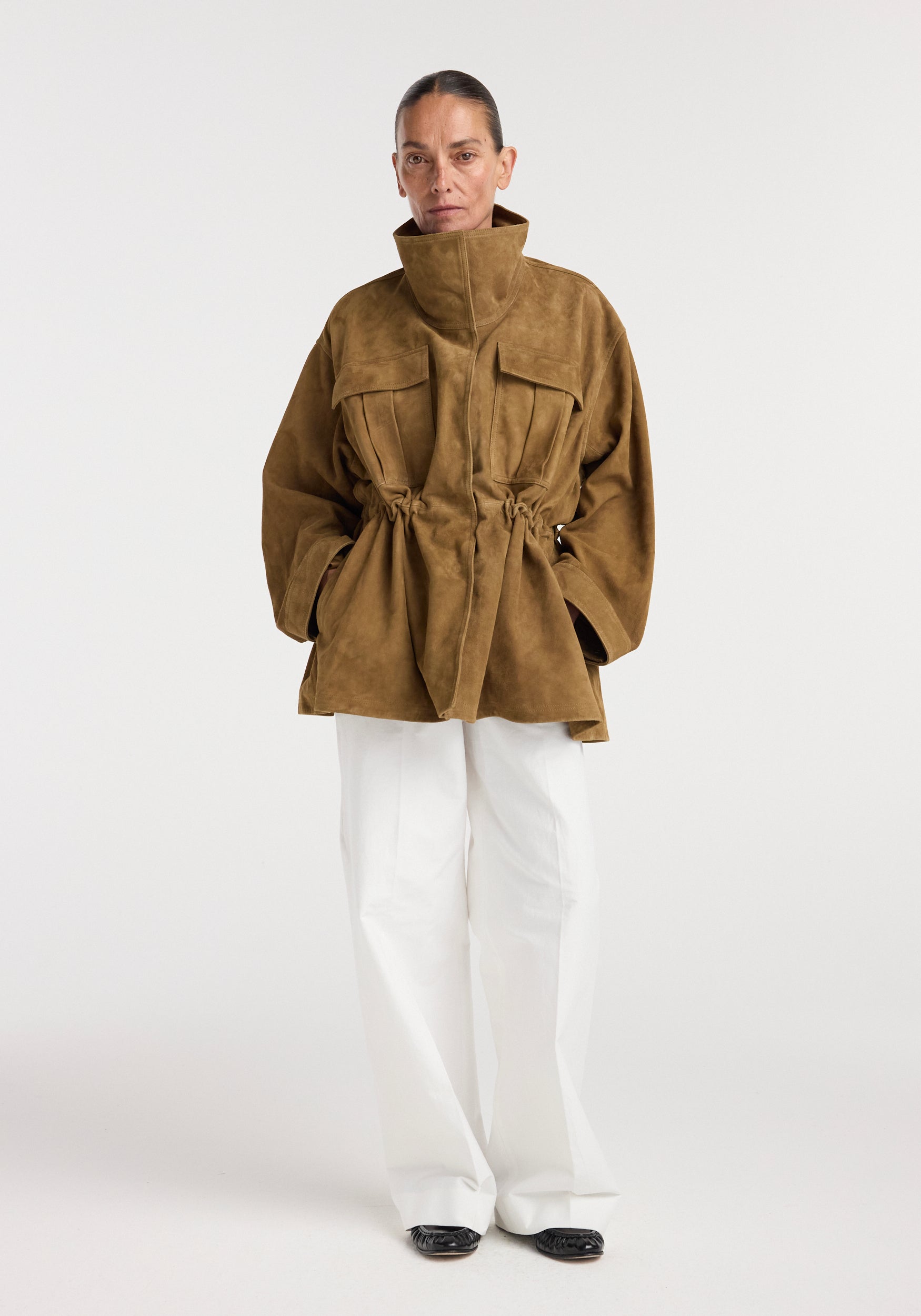 Model wearing a sculptural suede jacket | sage in a brownish shade with a high collar, cinched waist, and loose sleeves paired with white pants and black shoes against a plain background.