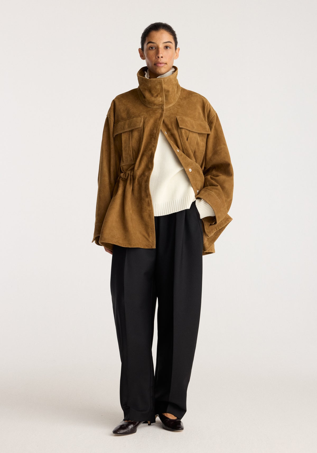 Sculptural suede jacket | sage worn by a model with a high collar and flap pockets over a cream sweater and black trousers