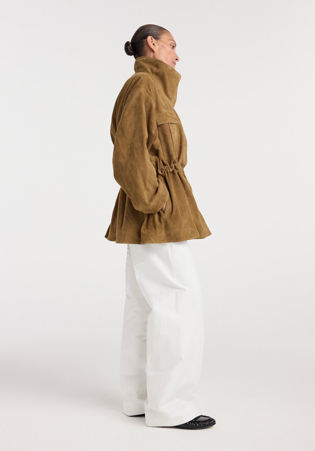 Woman standing in profile wearing a sculptural suede jacket | sage with a high collar and gathered waist, paired with wide white pants and black shoes against a plain white background.