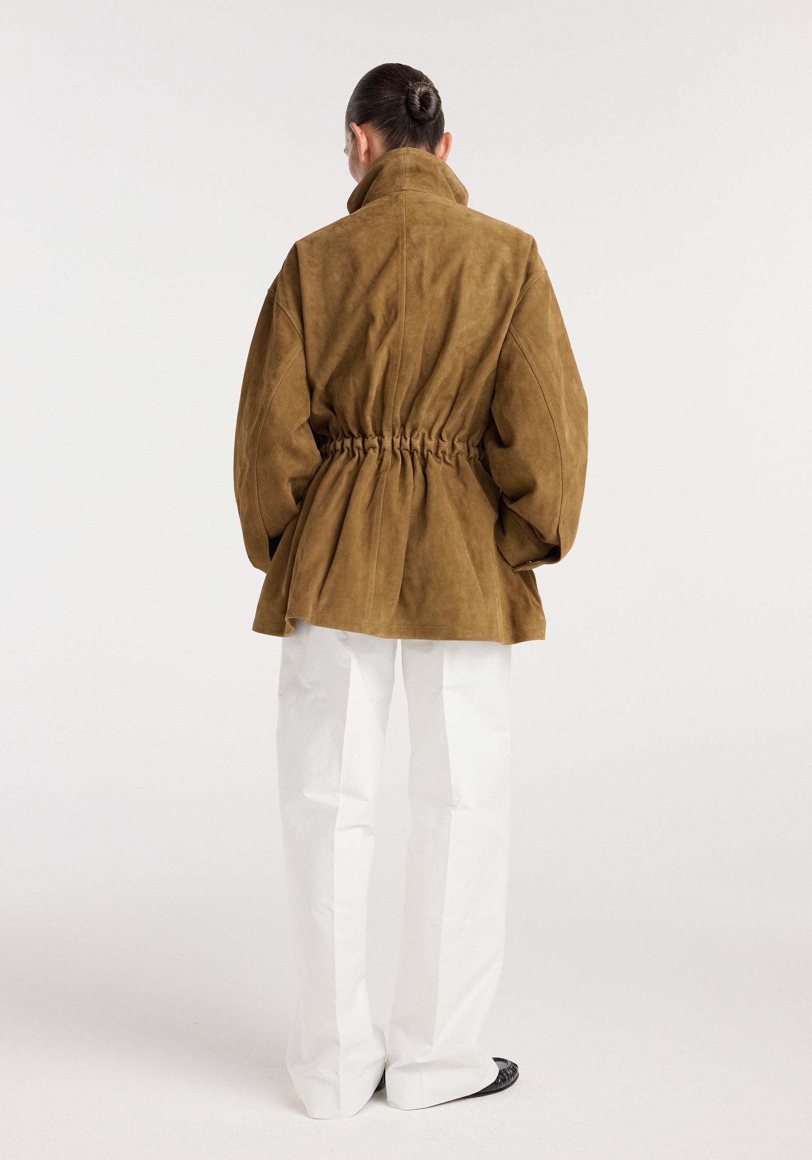 Back view of a model wearing a sculptural suede jacket | sage with gathered waist detail and loose sleeves, paired with white pants and black shoes against a plain background.