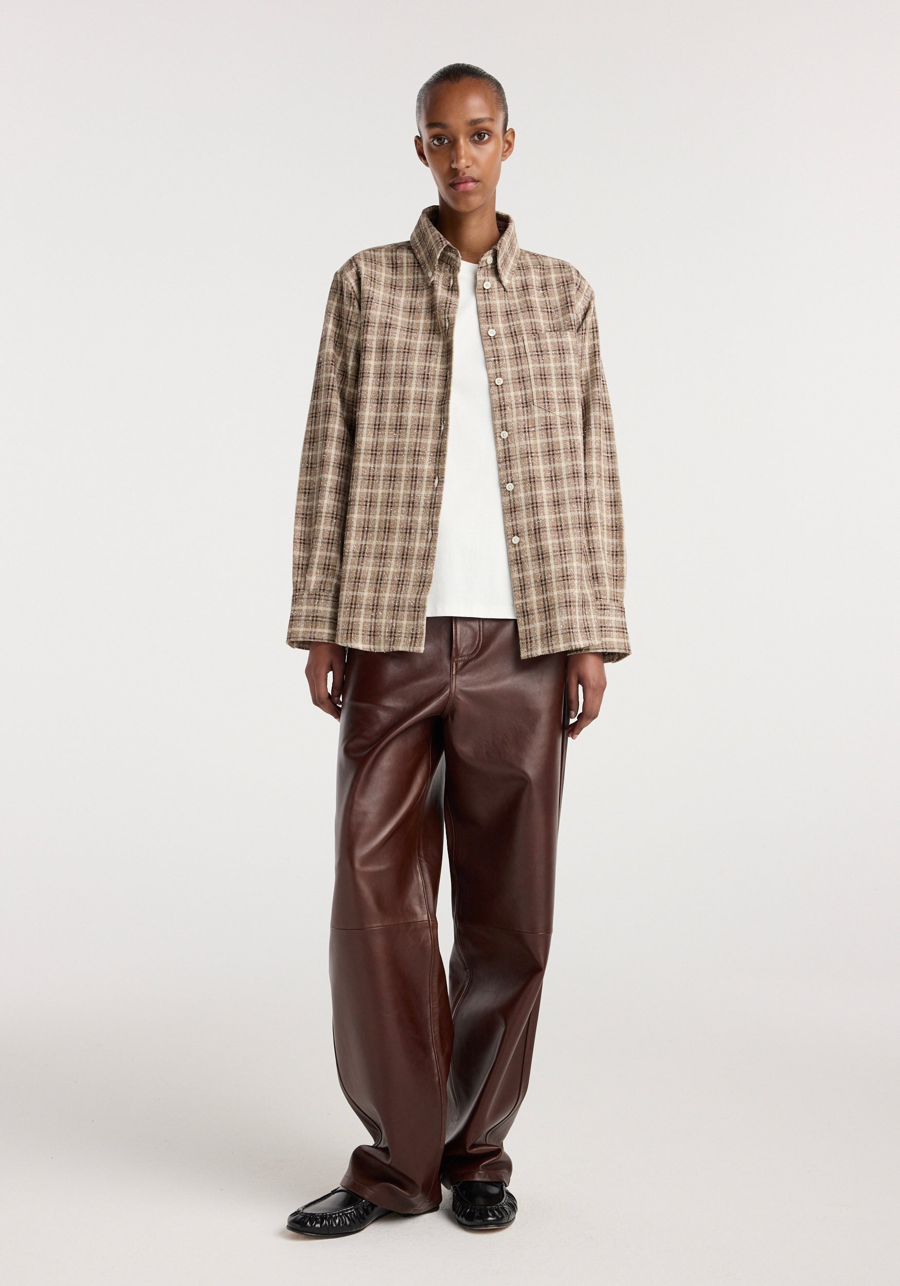 Model wearing an Oxford bouclé check shirt | boucle check in a neutral brown and beige check pattern, styled open over a white top with dark brown leather pants and black shoes.