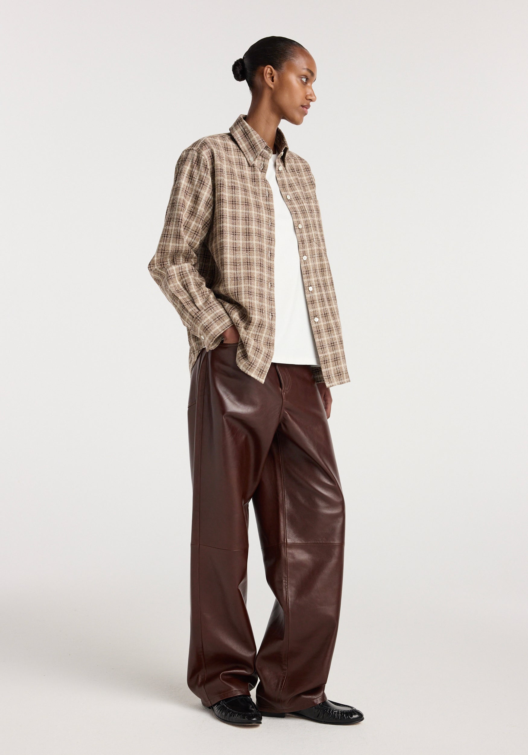 Model wearing an Oxford bouclé check shirt | boucle check in beige and brown tones over a white top, paired with wide-leg brown leather pants and black shoes, against a plain background.