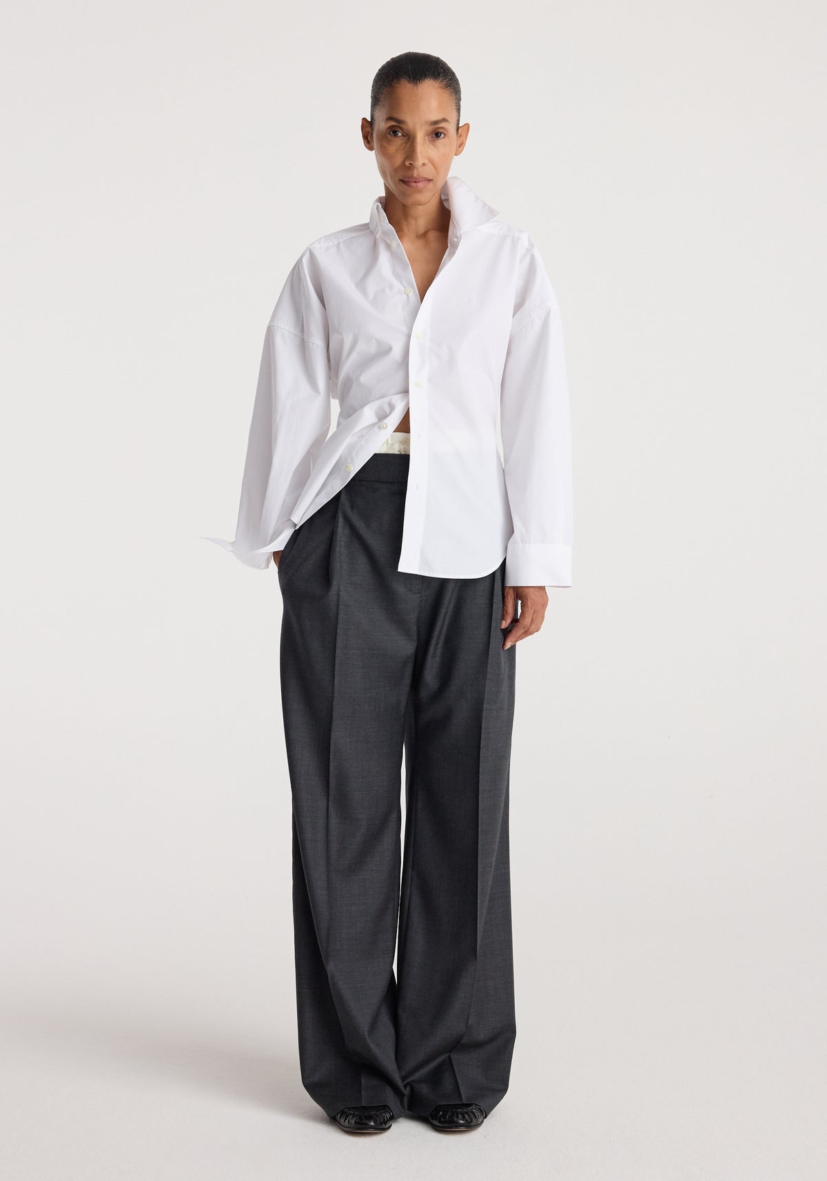 Model wearing a white Poplin uneven collar shirt | optic white with long sleeves and loose fit, paired with dark wide-leg trousers against a plain background.