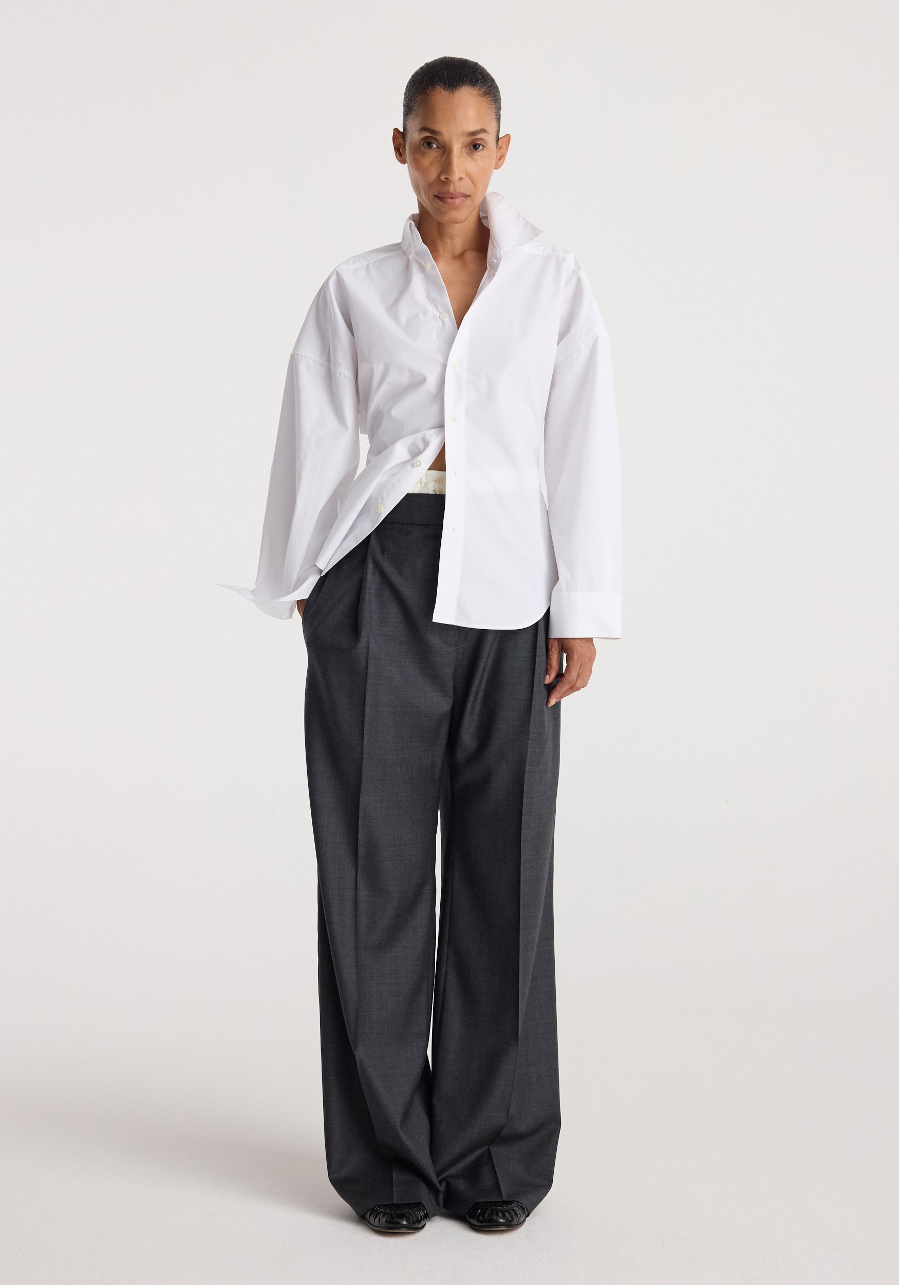 Model wearing a white Poplin uneven collar shirt | optic white with long sleeves and loose fit, paired with dark wide-leg trousers against a plain background.