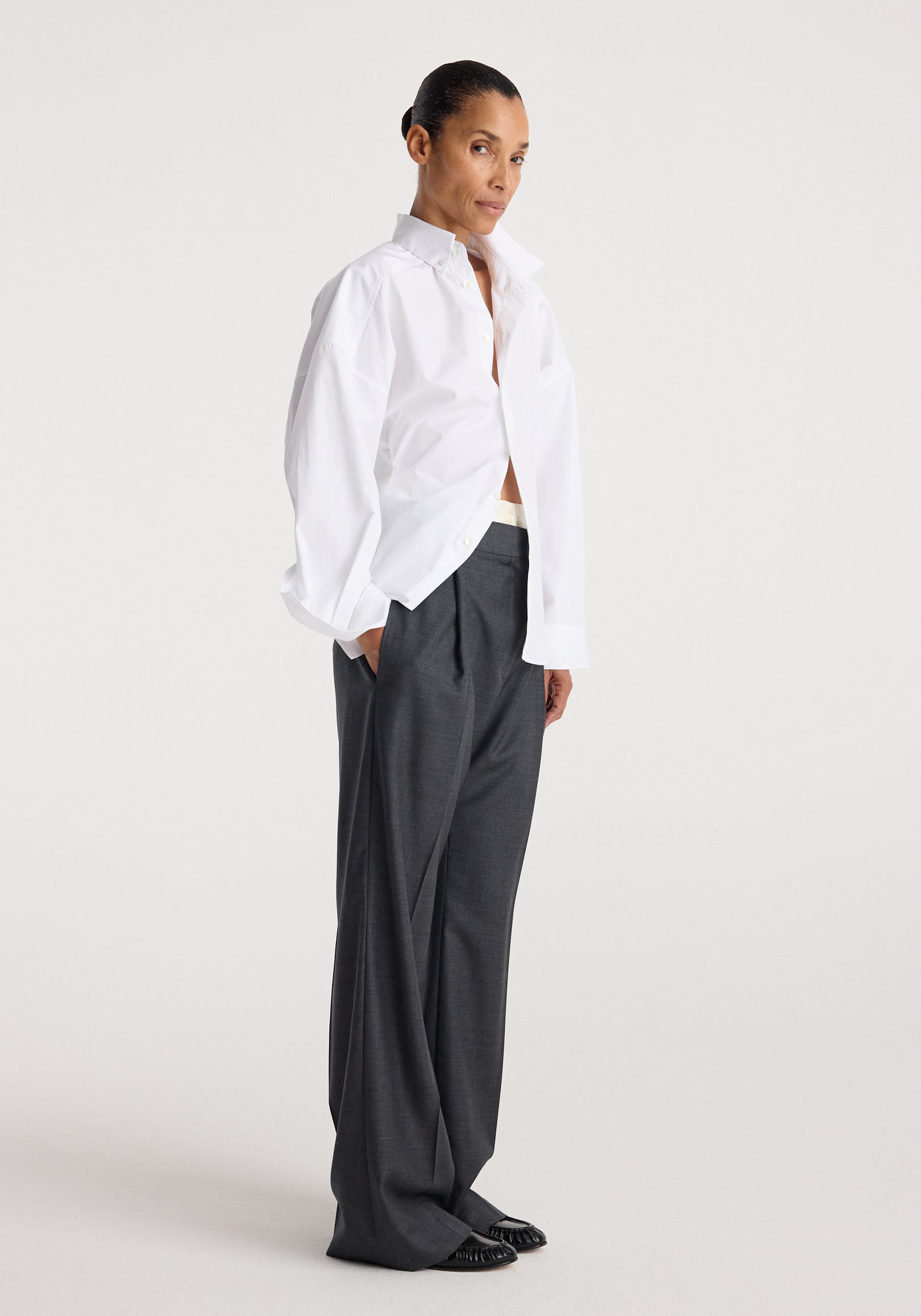 Woman wearing Poplin uneven collar shirt | optic white paired with loose dark gray trousers and black shoes, standing against a plain light background.