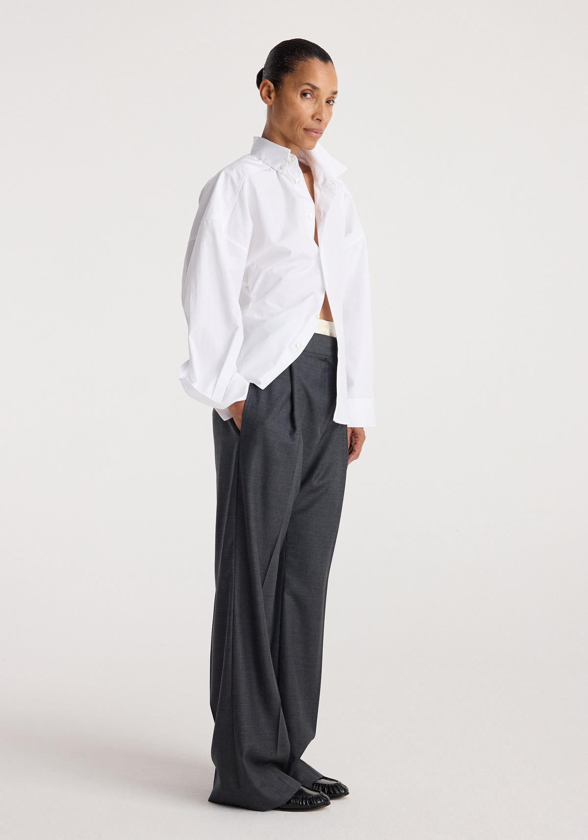Woman wearing Poplin uneven collar shirt | optic white paired with loose dark gray trousers and black shoes, standing against a plain light background.