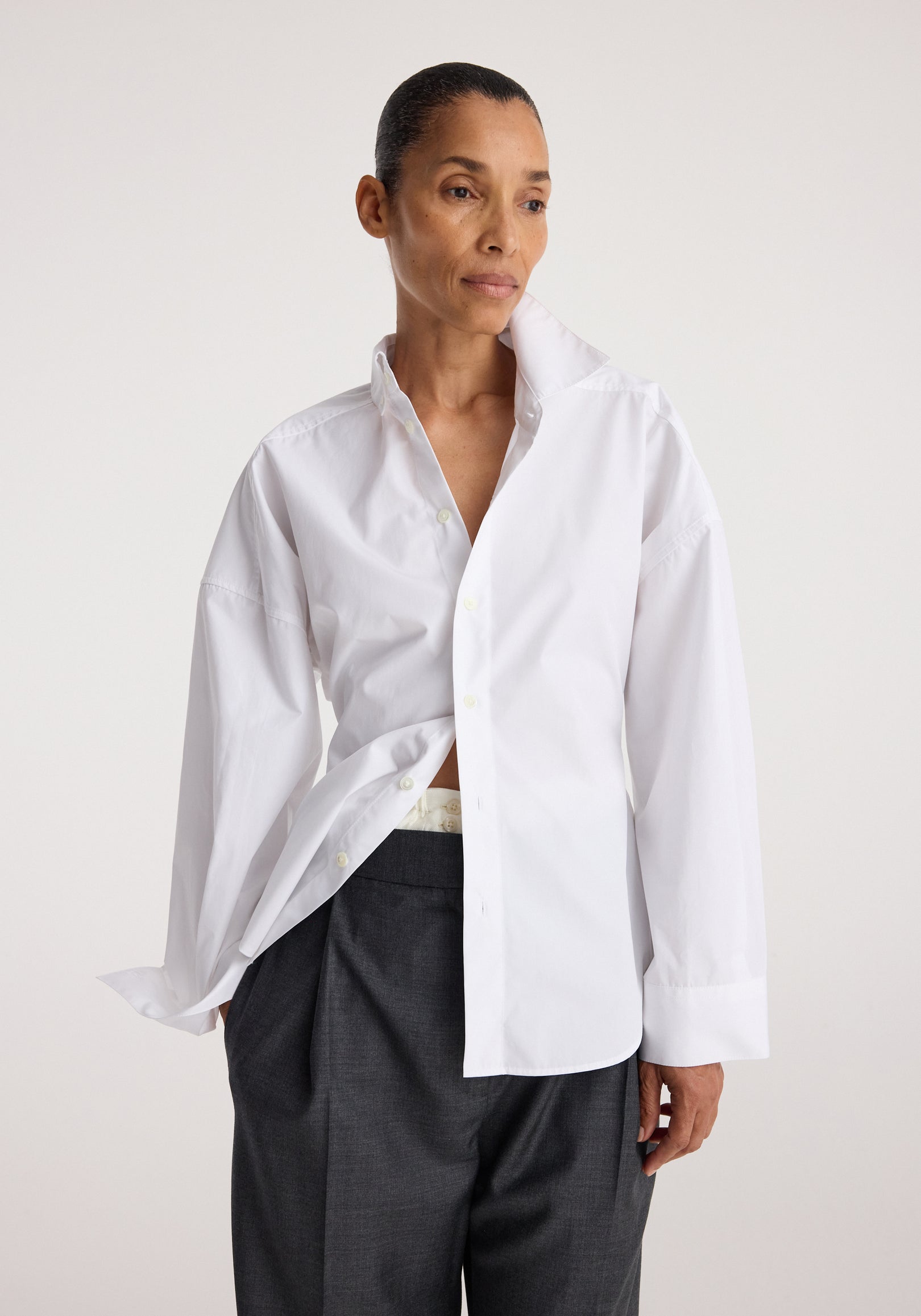 Woman wearing a white shirt with an uneven collar standing against a plain background, styled with dark gray pants, showcasing the Poplin uneven collar shirt | optic white.