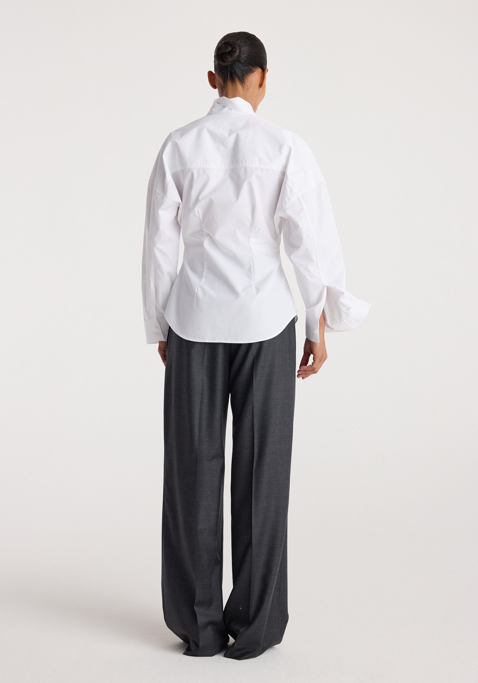 Back view of a person wearing a white Poplin uneven collar shirt | optic white paired with dark gray wide-leg trousers against a plain background.
