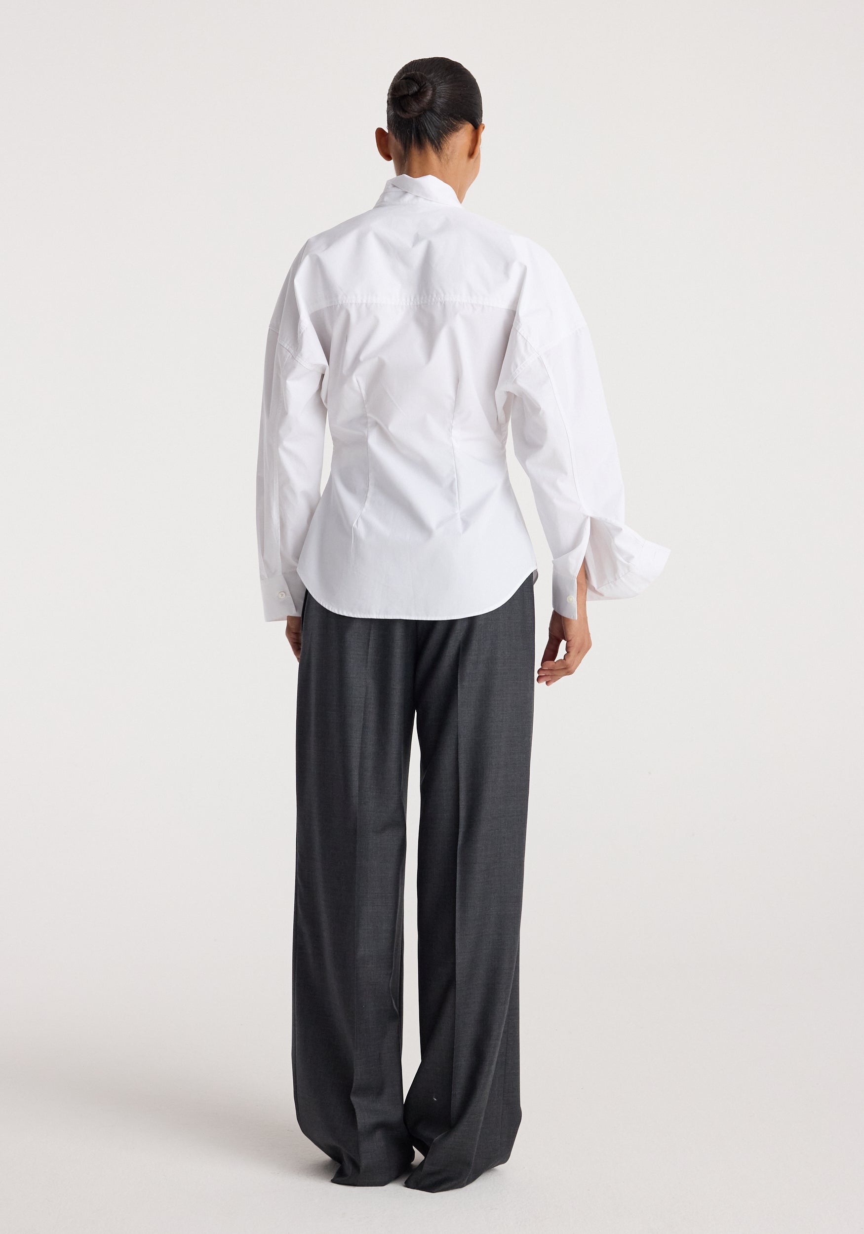 Back view of a person wearing a white Poplin uneven collar shirt | optic white paired with dark gray wide-leg trousers against a plain background.