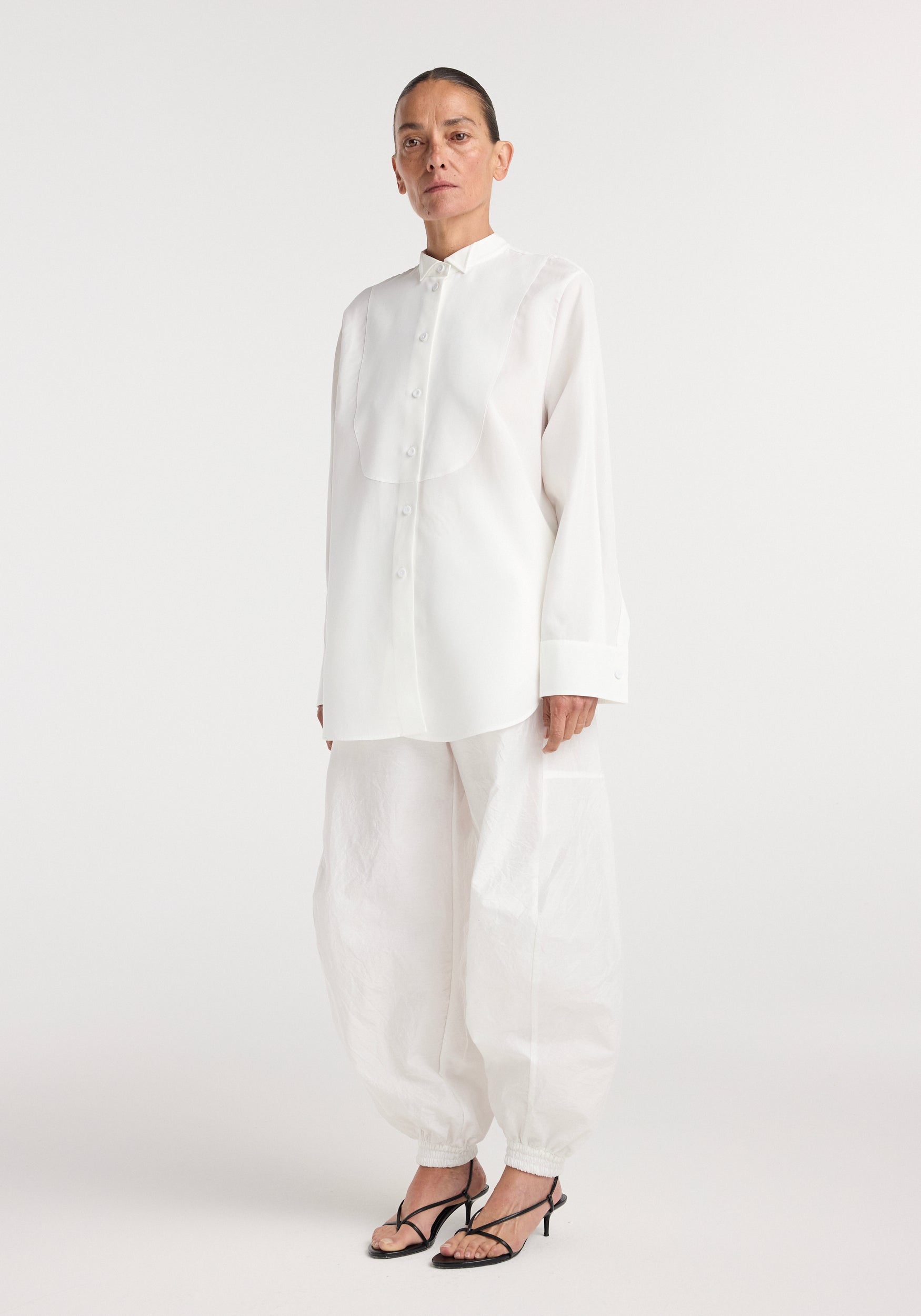 Model wearing a loose-fit tuxedo shirt | optic white with concealed buttons and classic cuffs, paired with matching white wide-leg pants and black strappy heels on a plain white background.