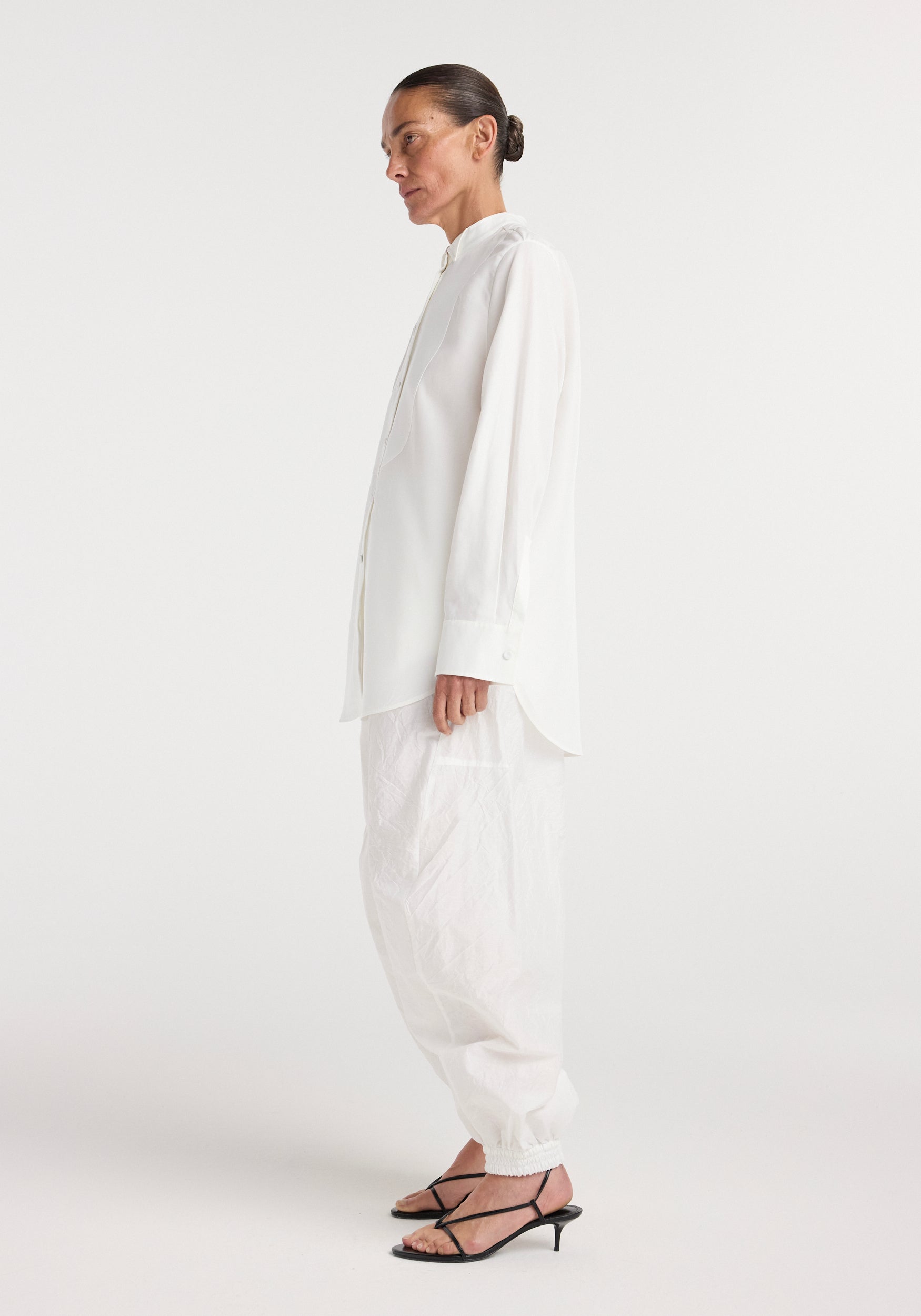 Side profile of a model wearing a formal Tuxedo shirt | optic white with a high collar and buttoned cuffs paired with loose white pants and black strappy heeled sandals on a plain background.