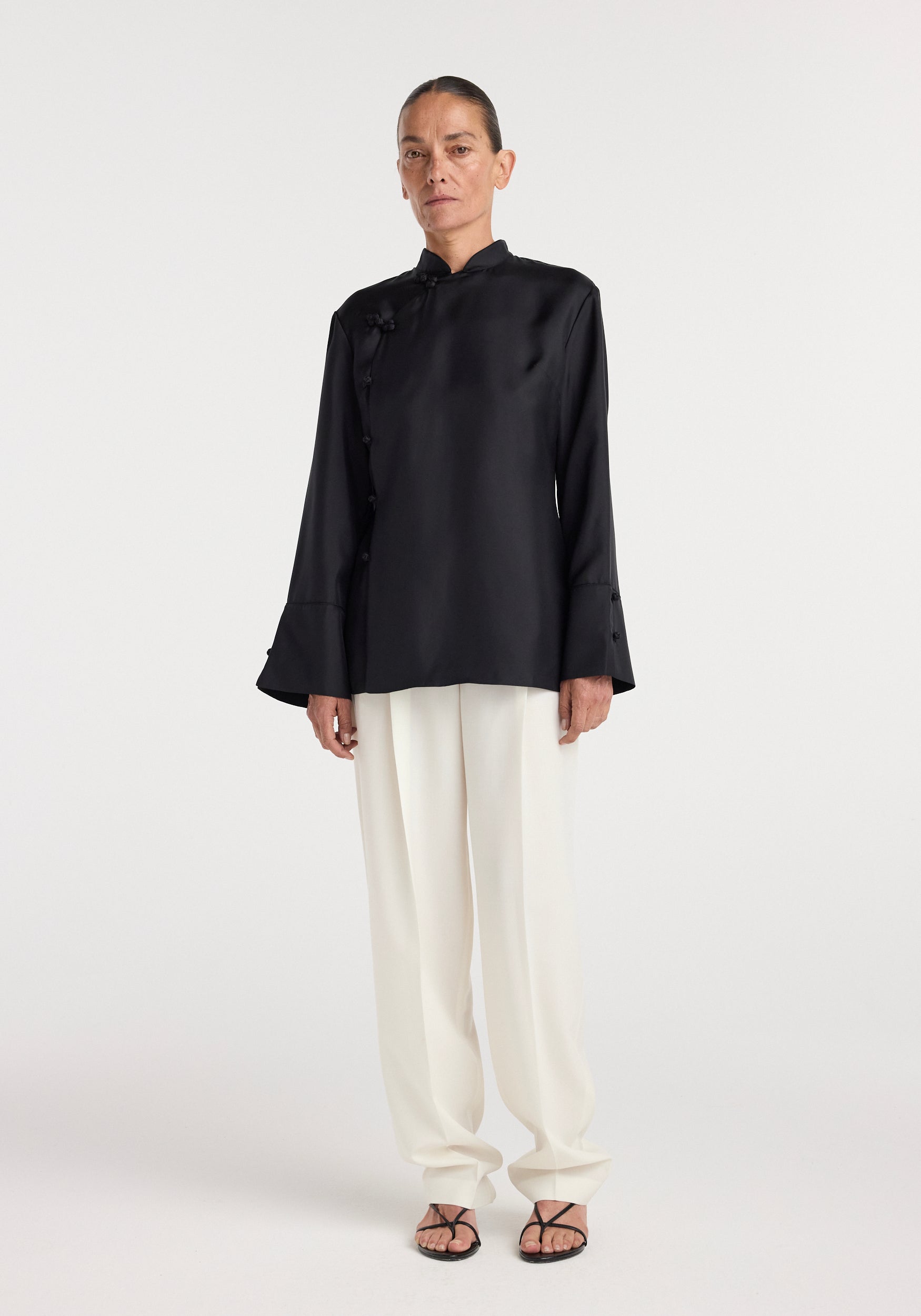 Woman wearing a black Pankou closure silk top | black with long sleeves and white loose trousers, standing against a plain light background.