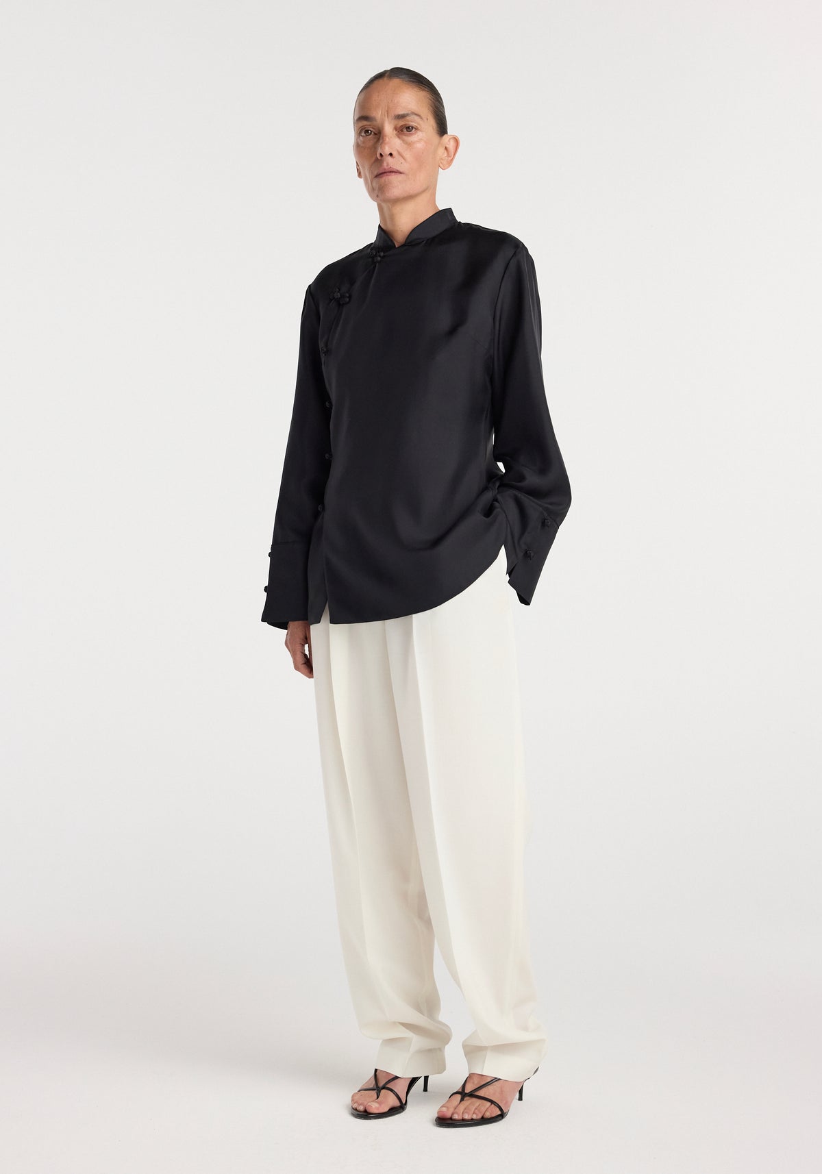 Woman wearing a black Pankou closure silk top | black with long sleeves and mandarin collar, paired with loose white pants and black strappy heels, standing against a plain background.