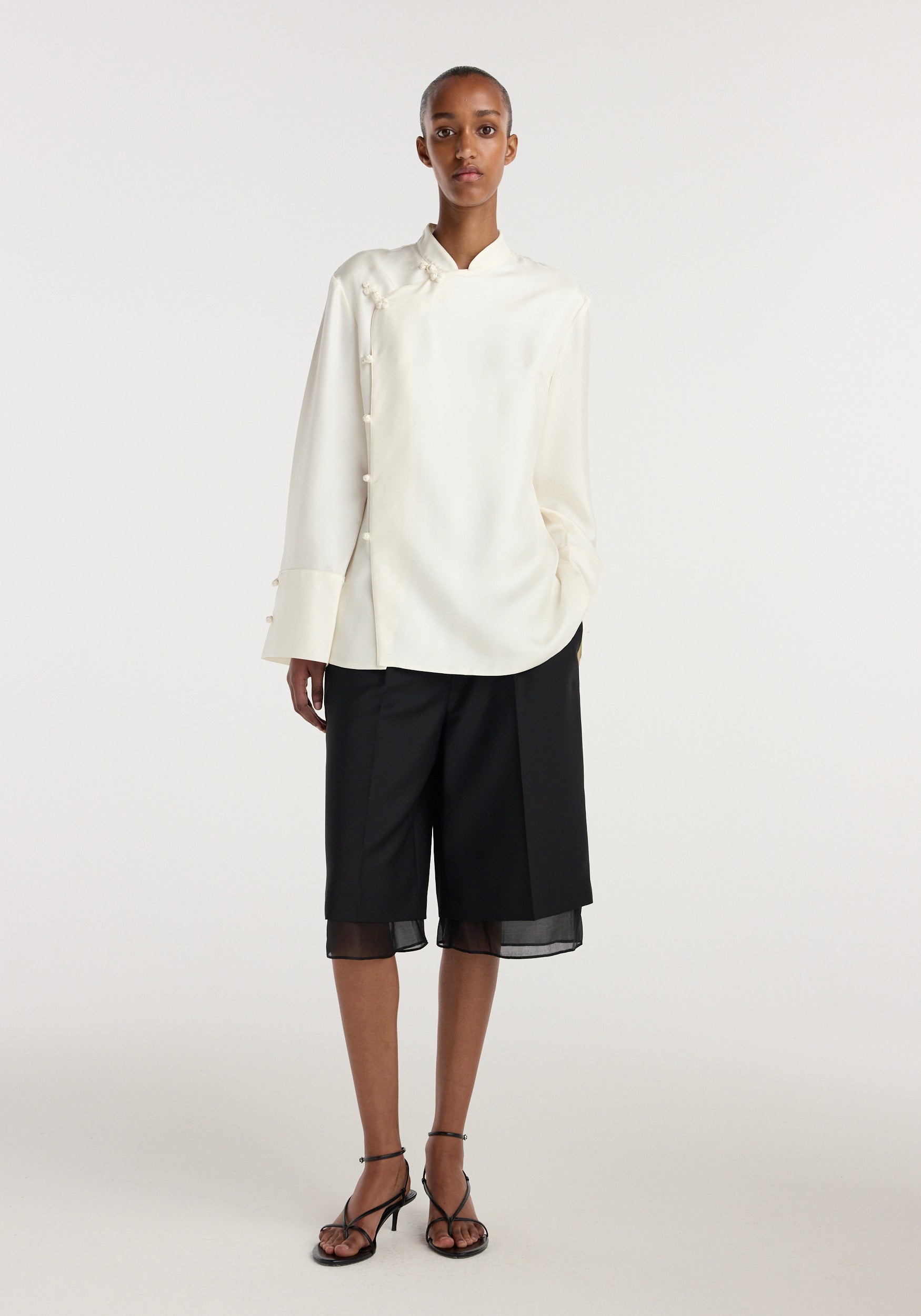 Model wearing a cream-colored Pankou closure silk top | cream with long sleeves and decorative button details paired with black knee-length shorts and black strappy heels.