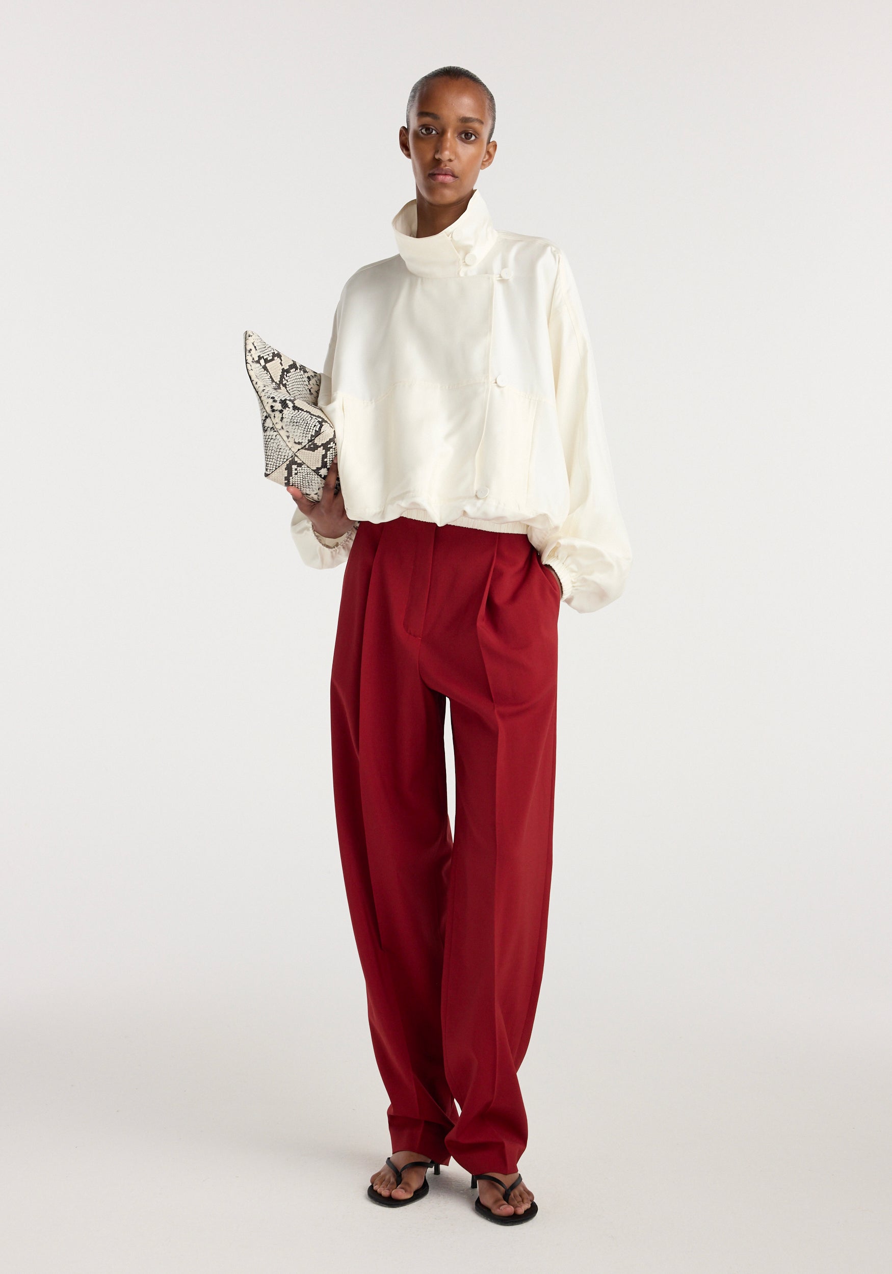 Model wearing a cream Silk blouson anorak with a high neck and loose fit, paired with wide-leg red trousers, holding a snakeskin-patterned clutch bag.