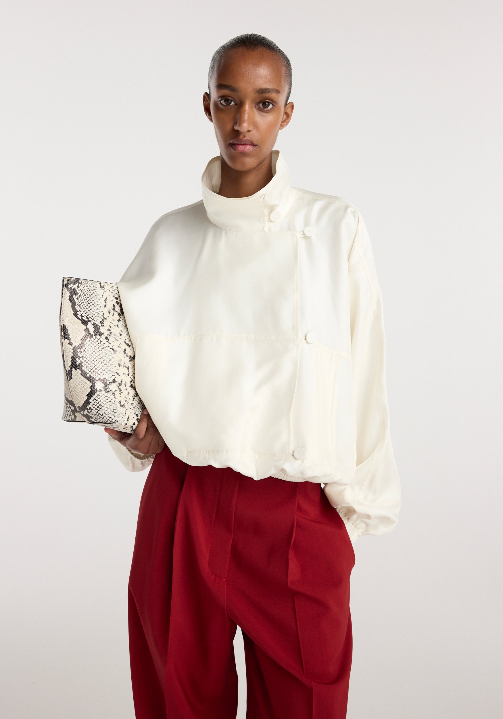 Model wearing a cream Silk blouson anorak with a high collar and buttons, paired with red trousers and holding a snake-print clutch bag.