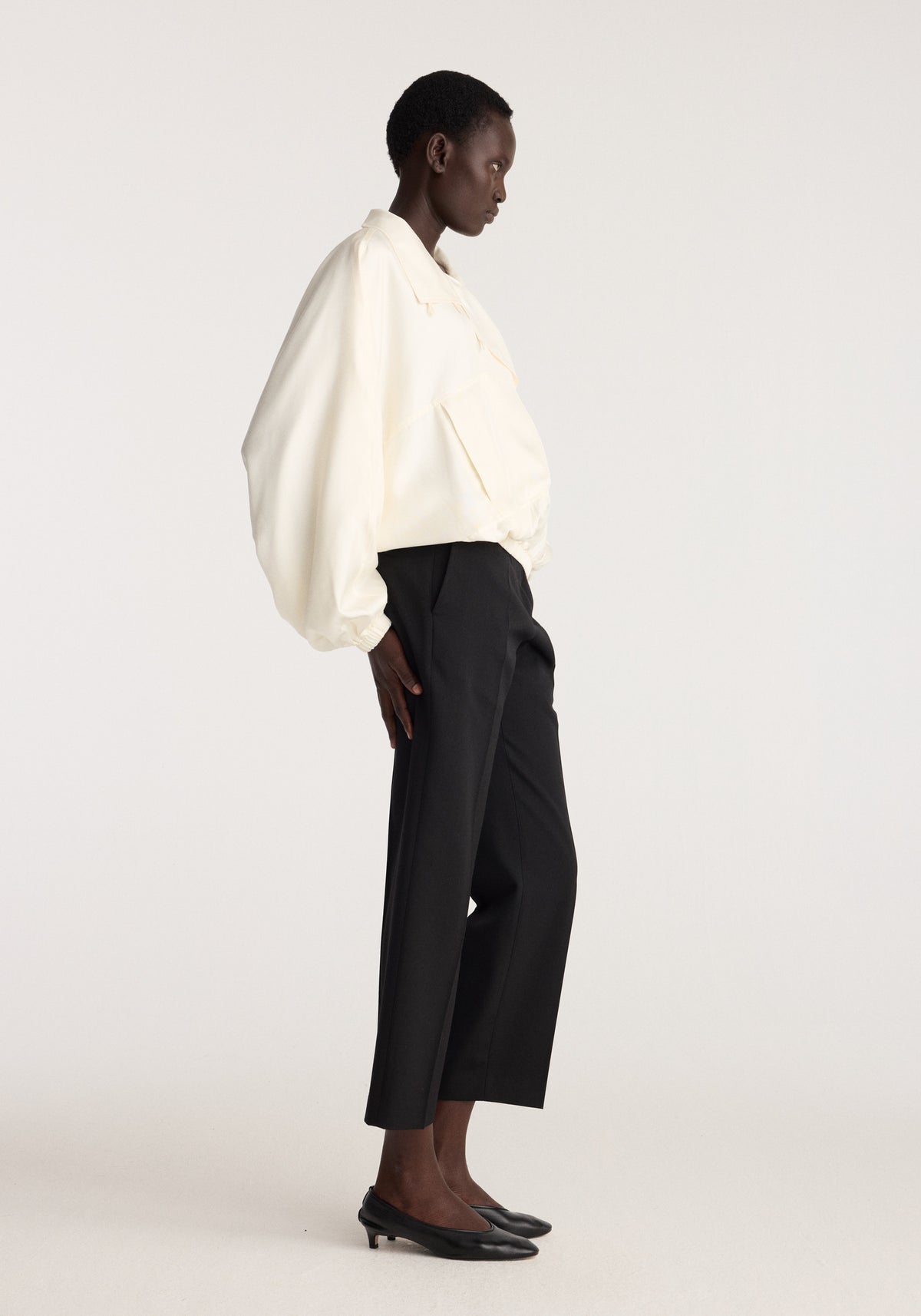 Model wearing Silk blouson anorak | cream with black cropped trousers and pointed flats in a side profile studio portrait