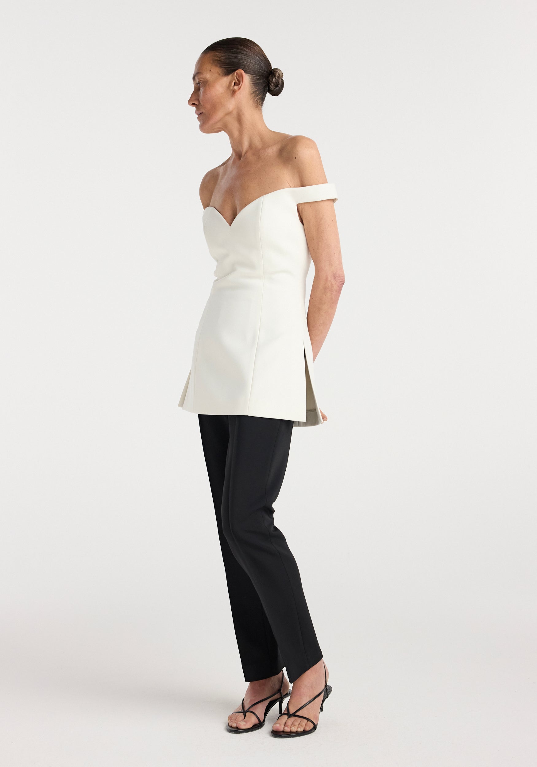 Woman wearing an off shoulder top | cream with a fitted bodice and side slits, paired with black slim pants and strappy heels, standing against a plain background.