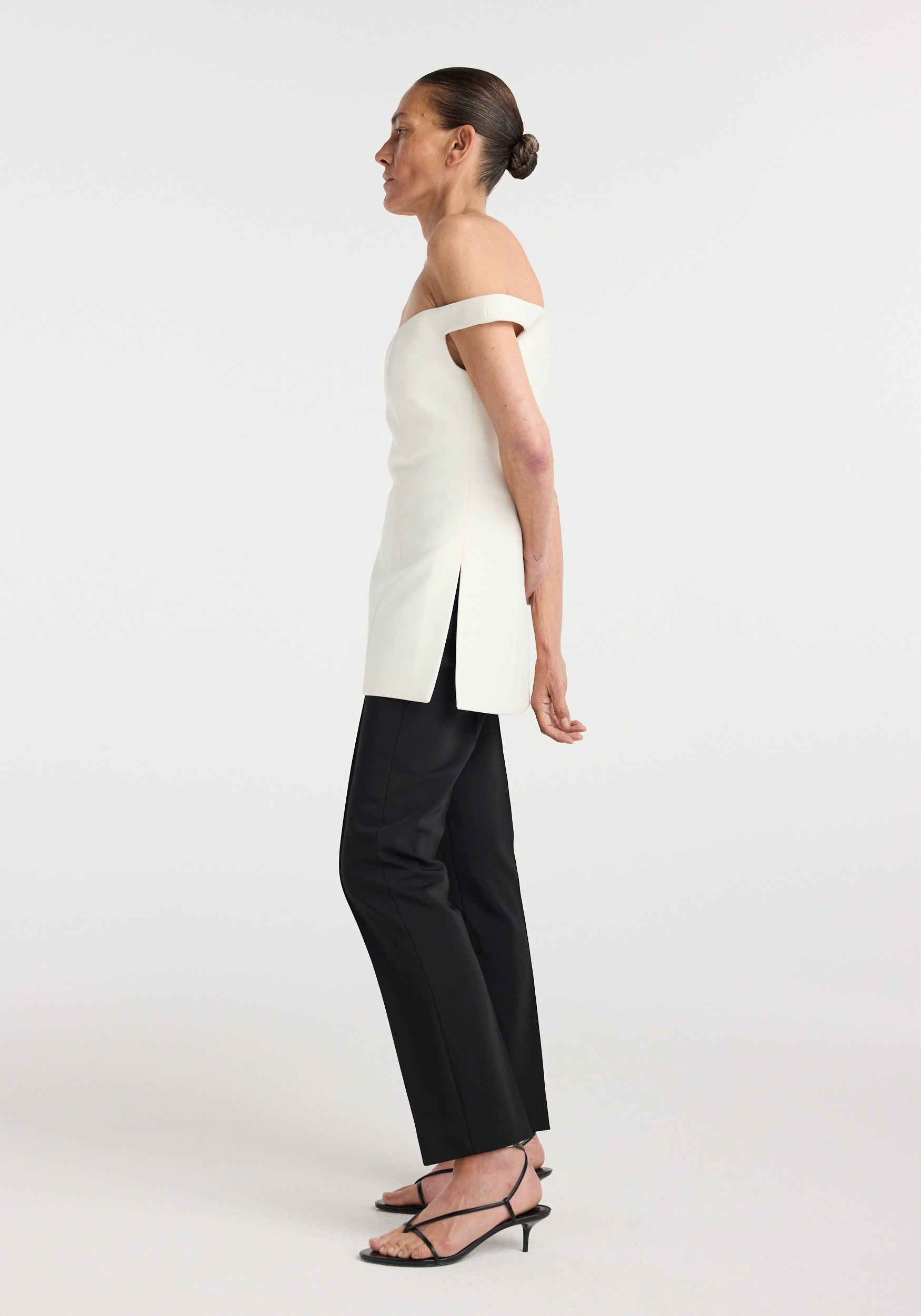 Woman wearing an off shoulder top | cream paired with black pants and strappy heeled sandals, standing in profile against a plain white background.