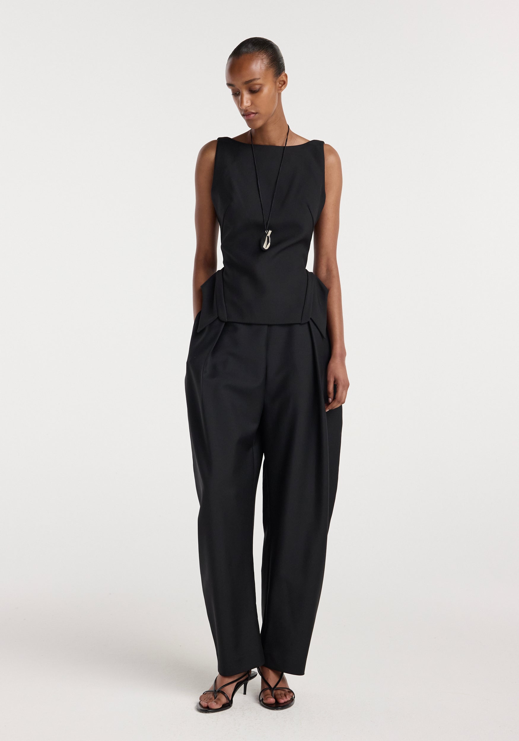 Woman wearing a sleeveless black top with a structured design paired with loose black trousers and black strappy heels, styled with a long pendant necklace, Deconstructed collar top | black.