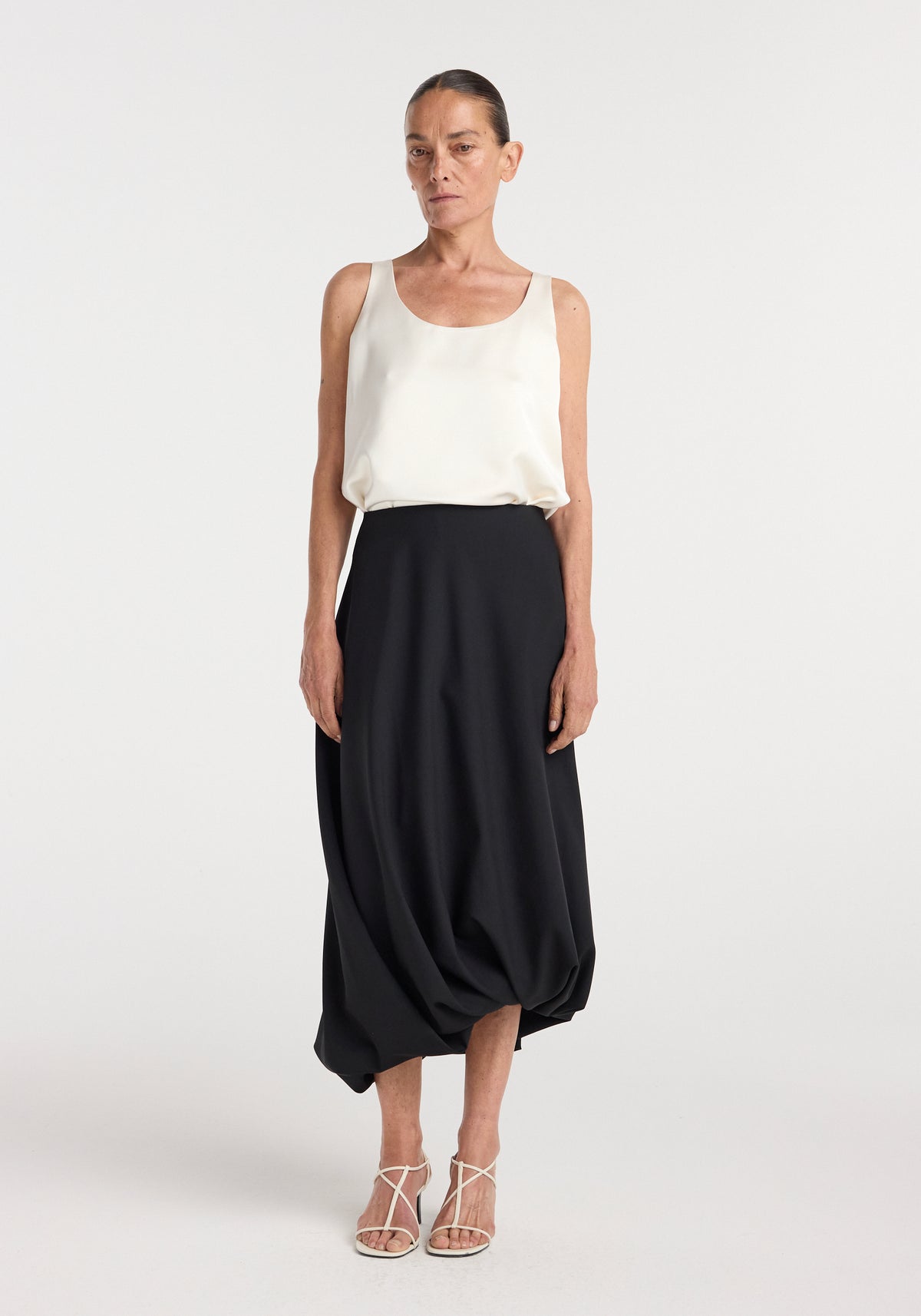 Woman wearing a white sleeveless top paired with a Twisted skirt | black, featuring a unique twisted hem detail, styled with white strappy heels against a plain background.