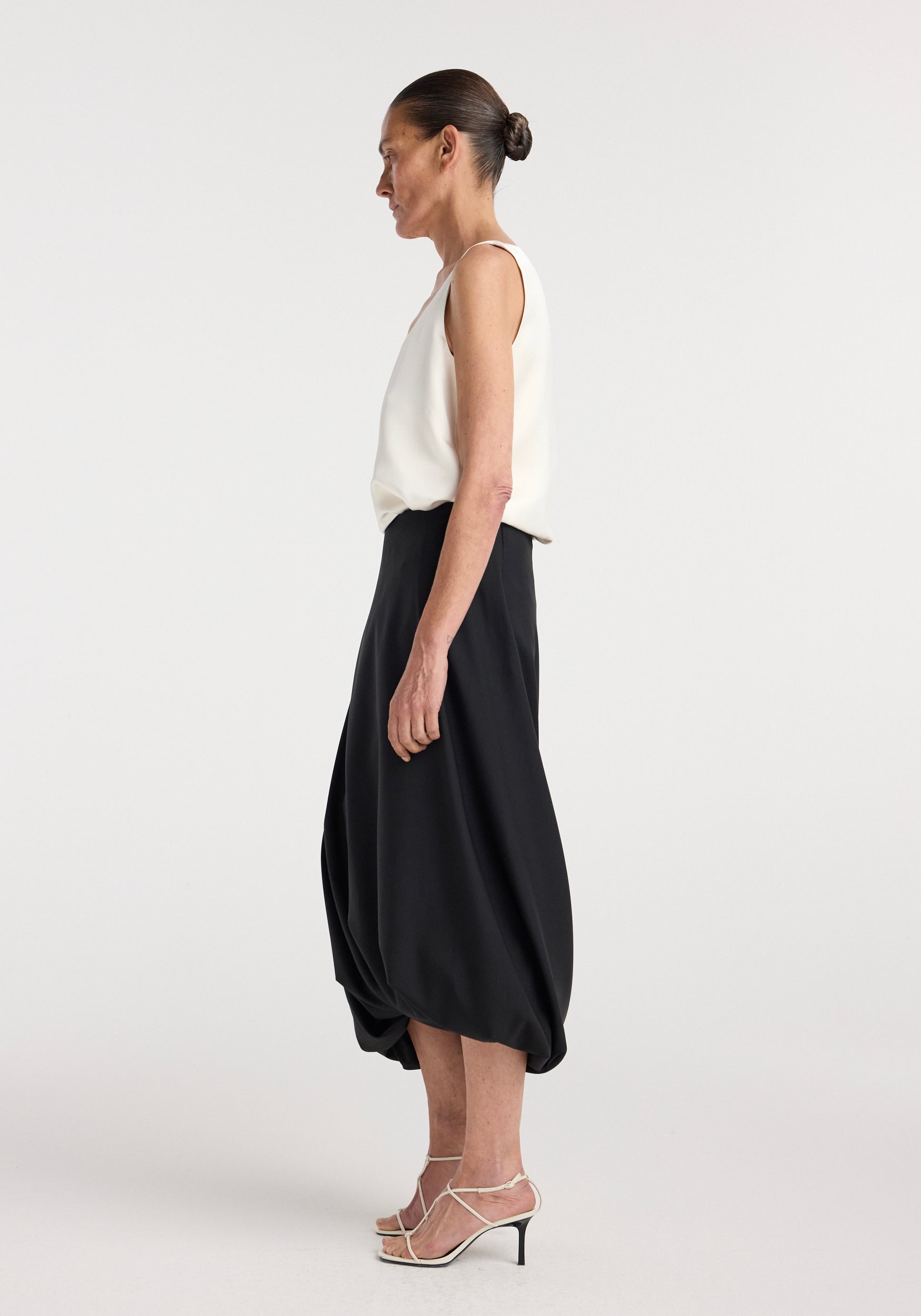 Woman wearing a satin scoop-neck top | ivory paired with a black midi skirt and strappy high-heeled sandals, shown in profile against a plain background.