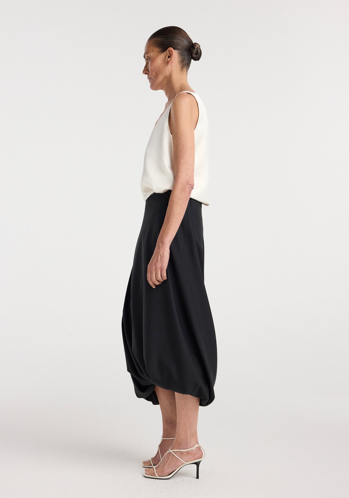 Profile view of a woman wearing a white sleeveless top paired with a black Twisted skirt | black featuring a gathered, twisted hem, styled with strappy white high-heeled sandals.