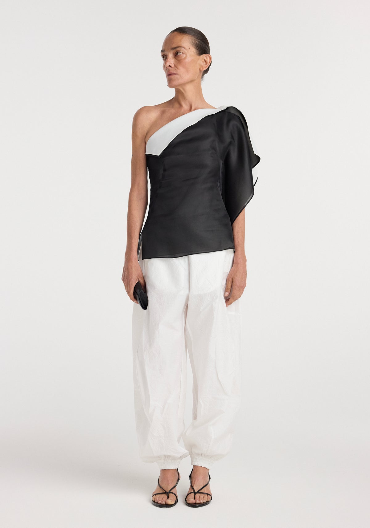 Woman wearing a black and white sculptural double layered top | black with asymmetrical one-shoulder design, paired with loose white pants and black sandals.