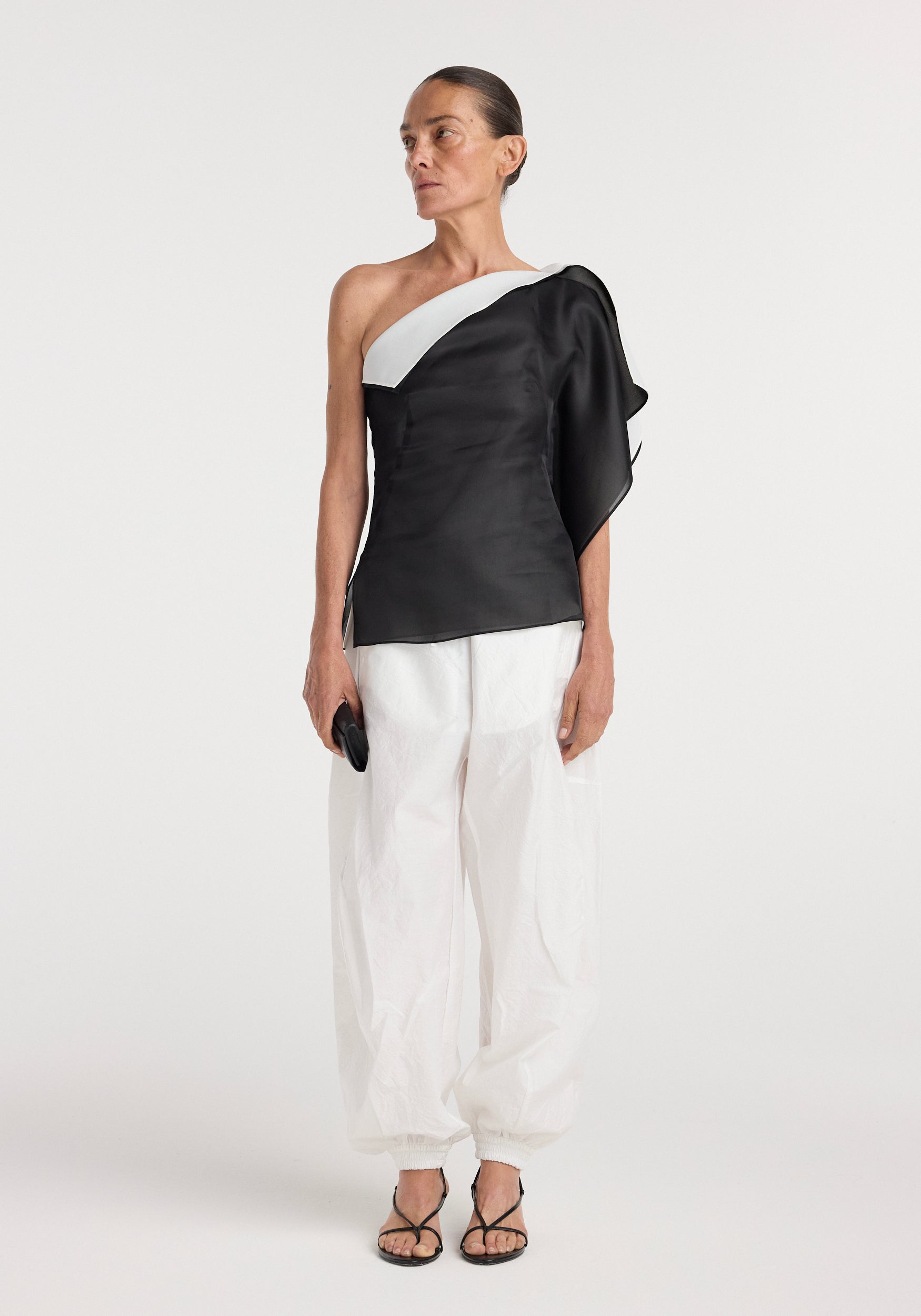 Woman wearing a black and white sculptural double layered top | black with asymmetrical one-shoulder design, paired with loose white pants and black sandals.