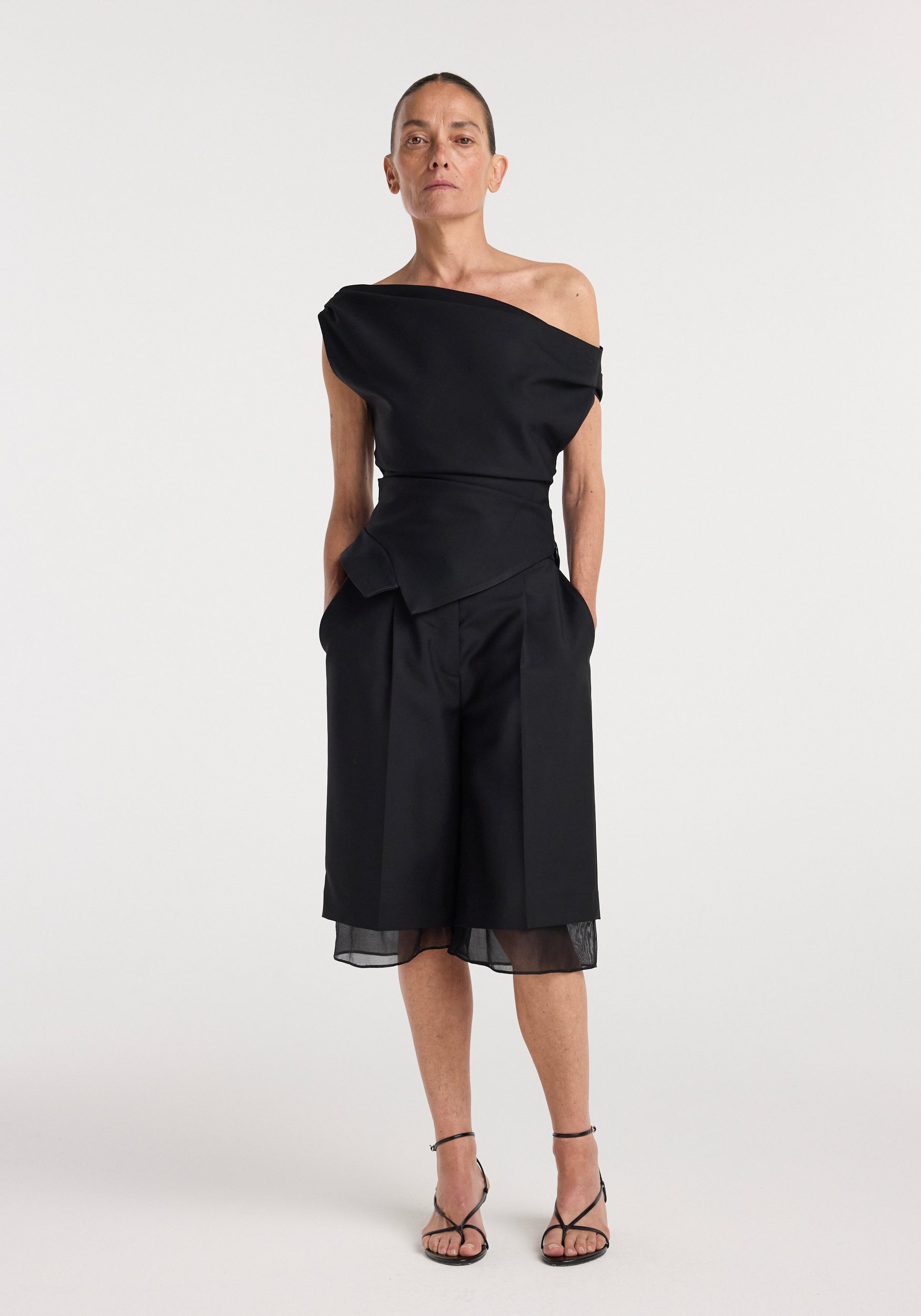 Woman wearing an off shoulder lapel top | black paired with matching black wide-leg shorts with sheer hems and black strappy heels, standing against a plain white background.