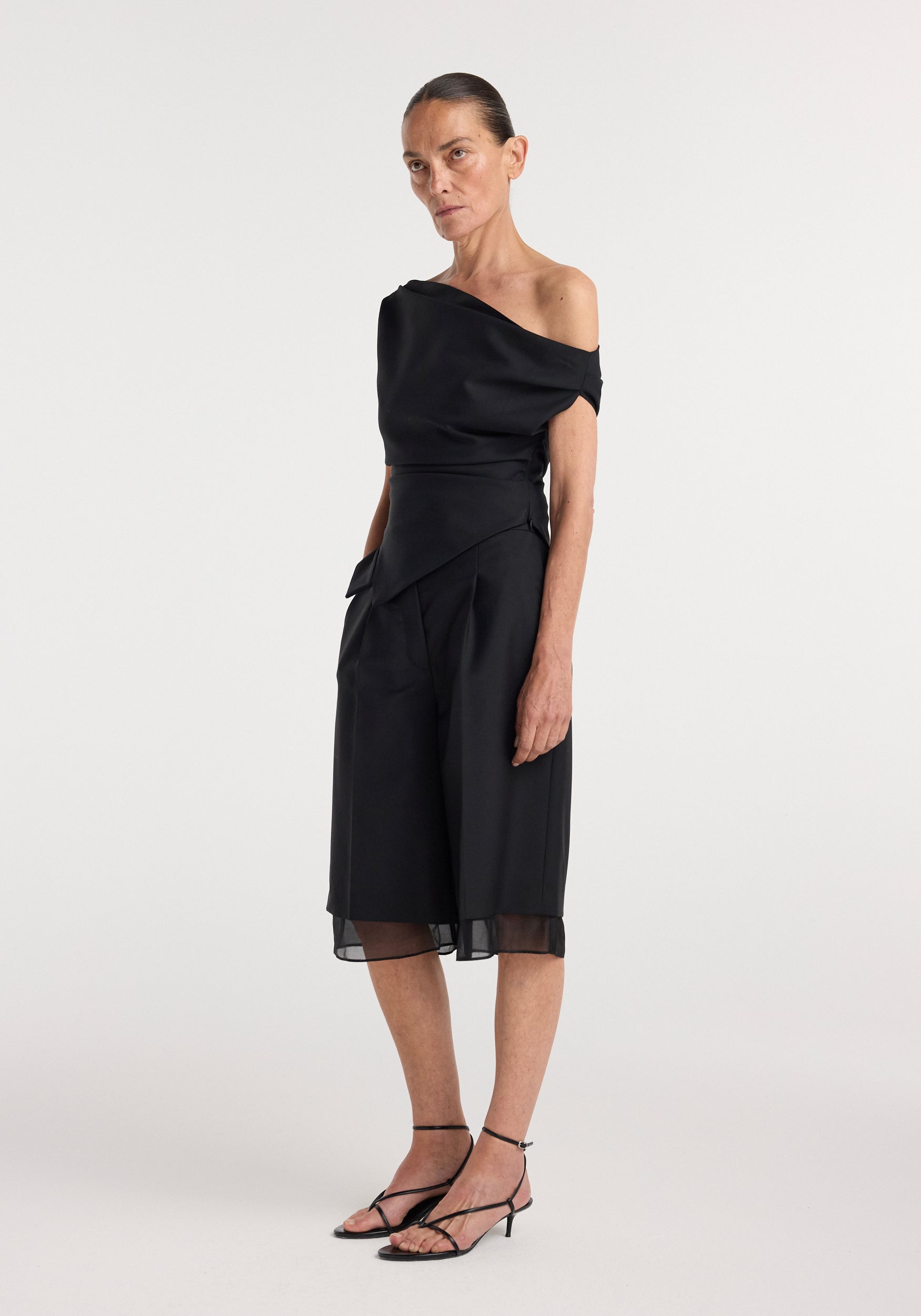 Woman wearing black off shoulder lapel top | black paired with wide-leg cropped pants and strappy black heels against a plain white background.