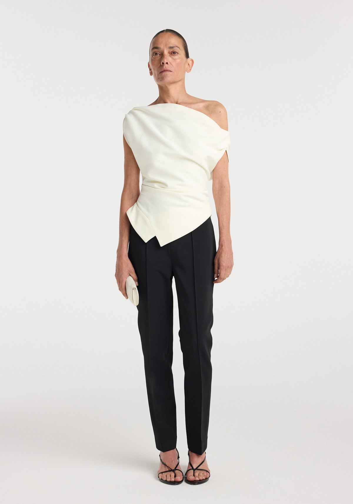 Woman wearing an off shoulder lapel top | off white styled with black tailored pants, holding a small clutch and wearing strappy black heels against a white background.