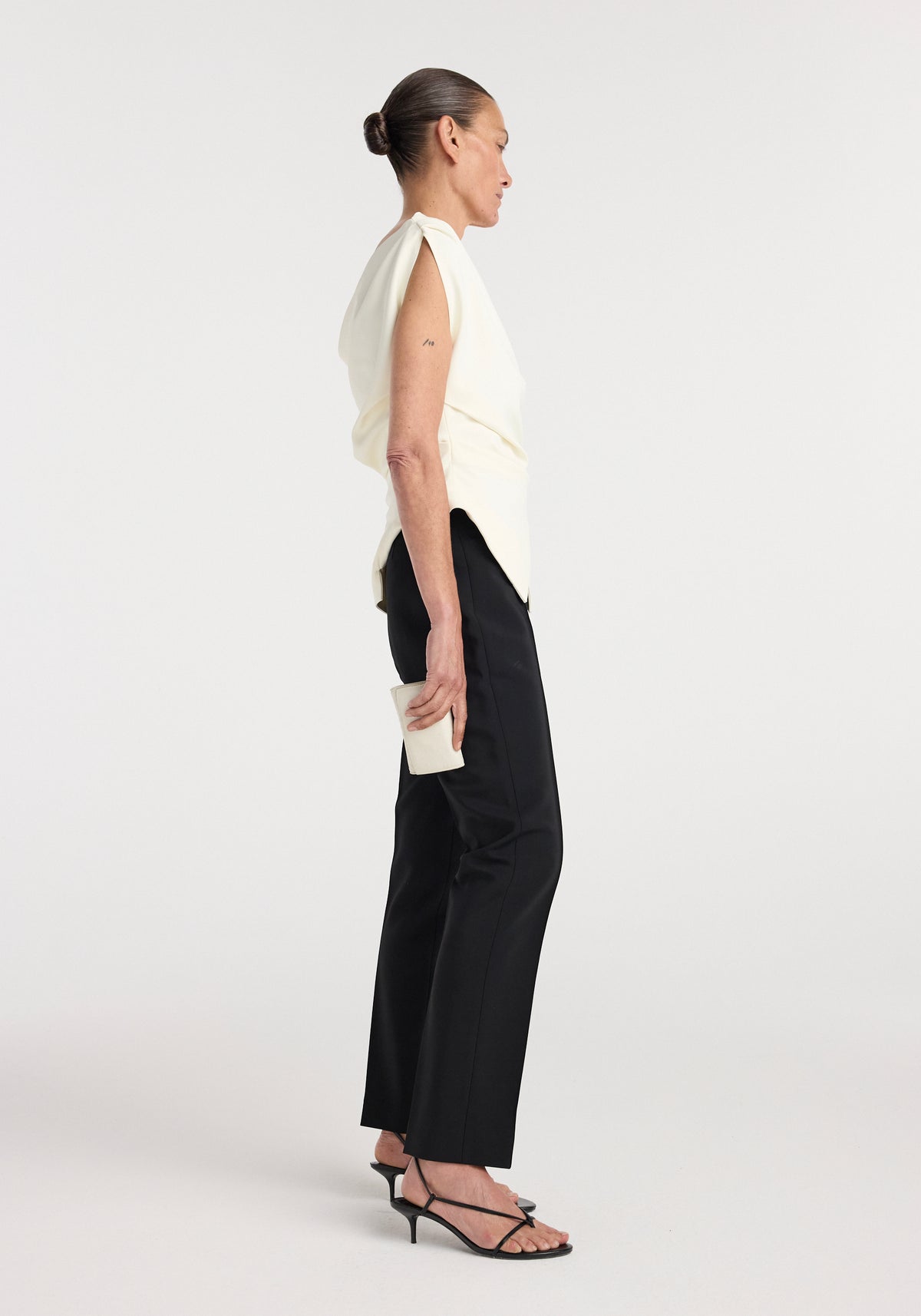 Side view of a woman wearing an off shoulder lapel top | off white paired with black pants and black heeled sandals, holding a small white clutch against a plain background.
