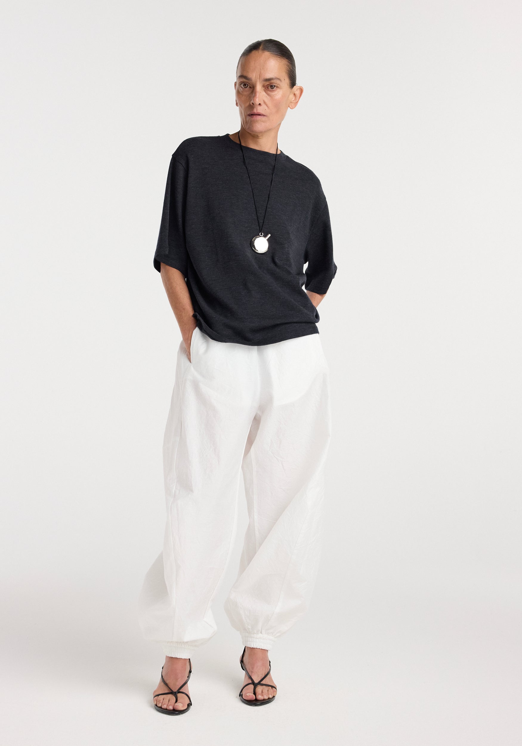 Woman wearing an oversized merino t-shirt | anthracite melange with loose white pants and black strappy sandals, standing against a plain light background.