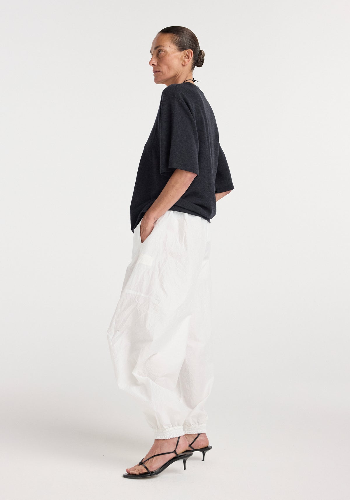 Woman wearing an oversized merino t-shirt | anthracite melange with loose white pants and black strappy heels, standing in profile against a plain background.