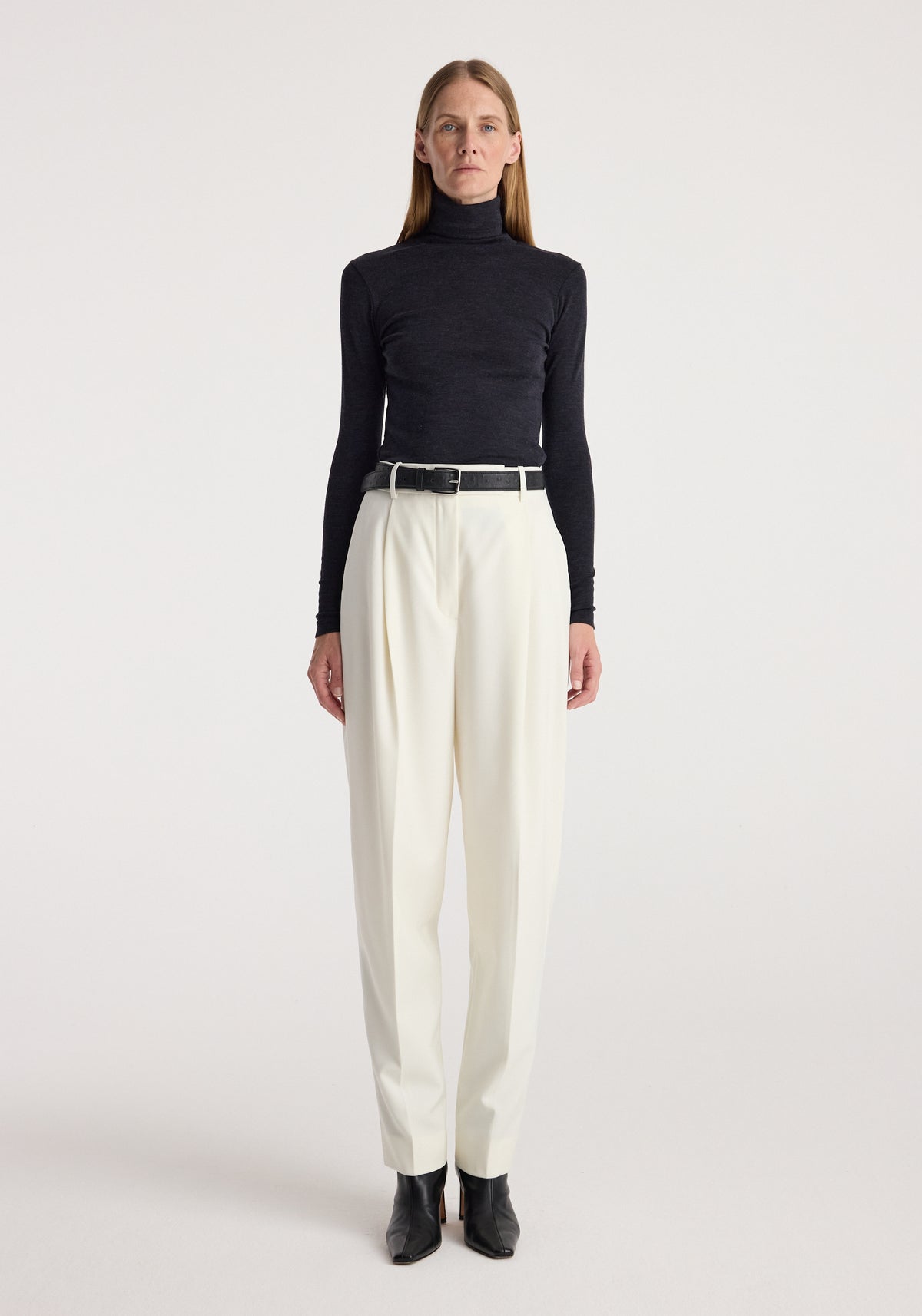 Woman wearing a black Fine merino turtleneck | anthracite melange paired with white high-waisted trousers, a black belt, and black ankle boots standing against a plain background.