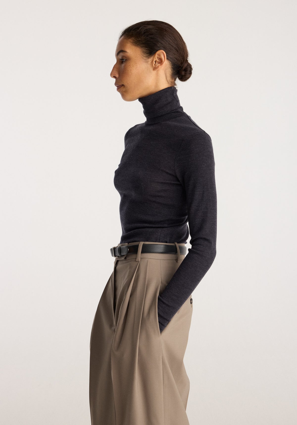 Fine merino turtleneck | anthracite melange worn by a model in profile with tailored taupe trousers and hands in pockets