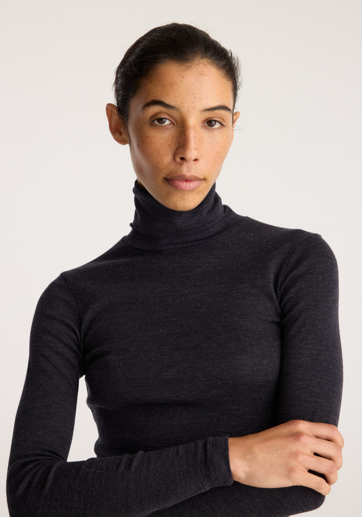 Model wearing Fine merino turtleneck | anthracite melange in a fitted dark charcoal knit with long sleeves and a high neck posed with folded arms
