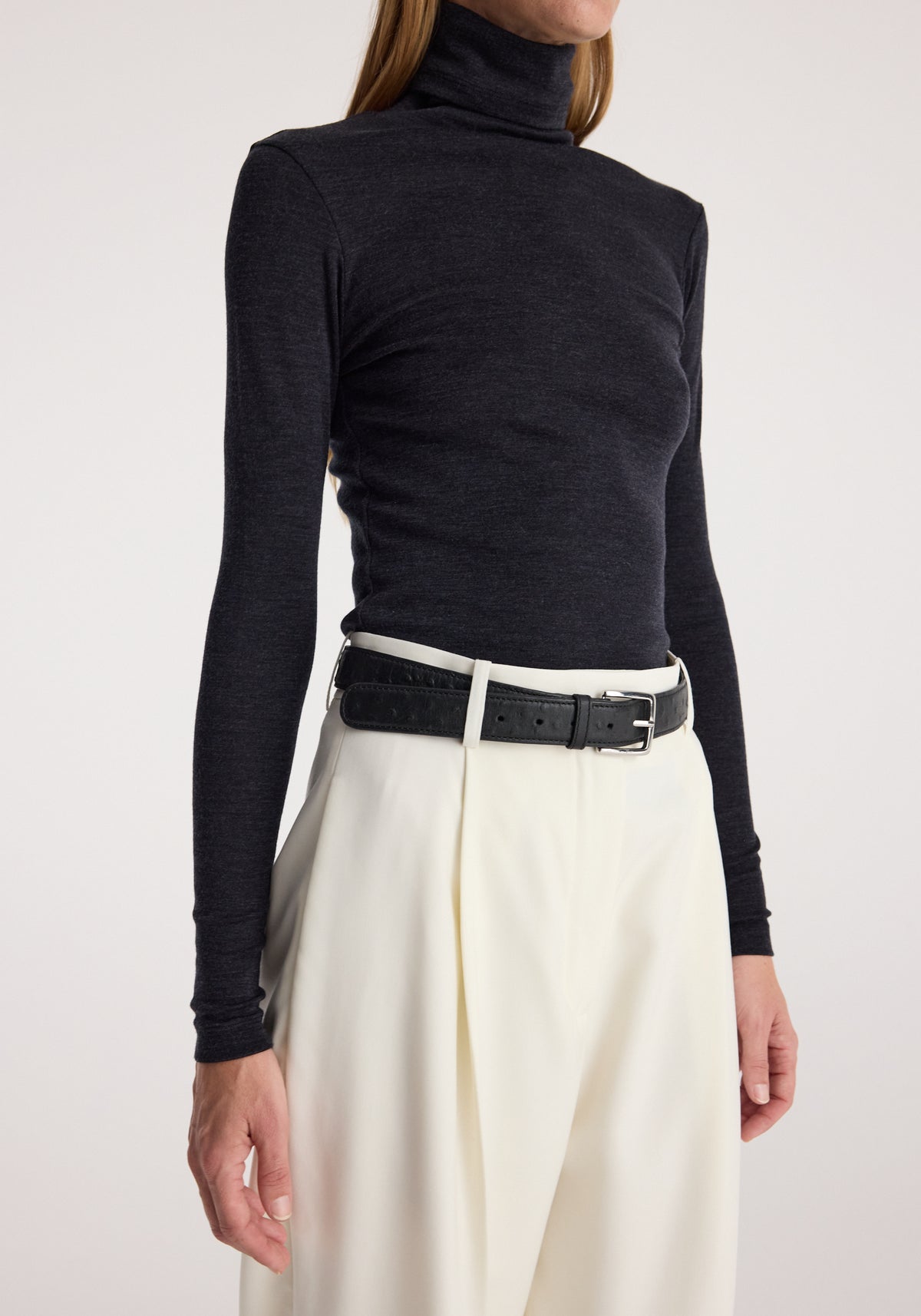Woman wearing a black Fine merino turtleneck | anthracite melange tucked into high-waisted cream trousers with a black belt, posing against a plain background.