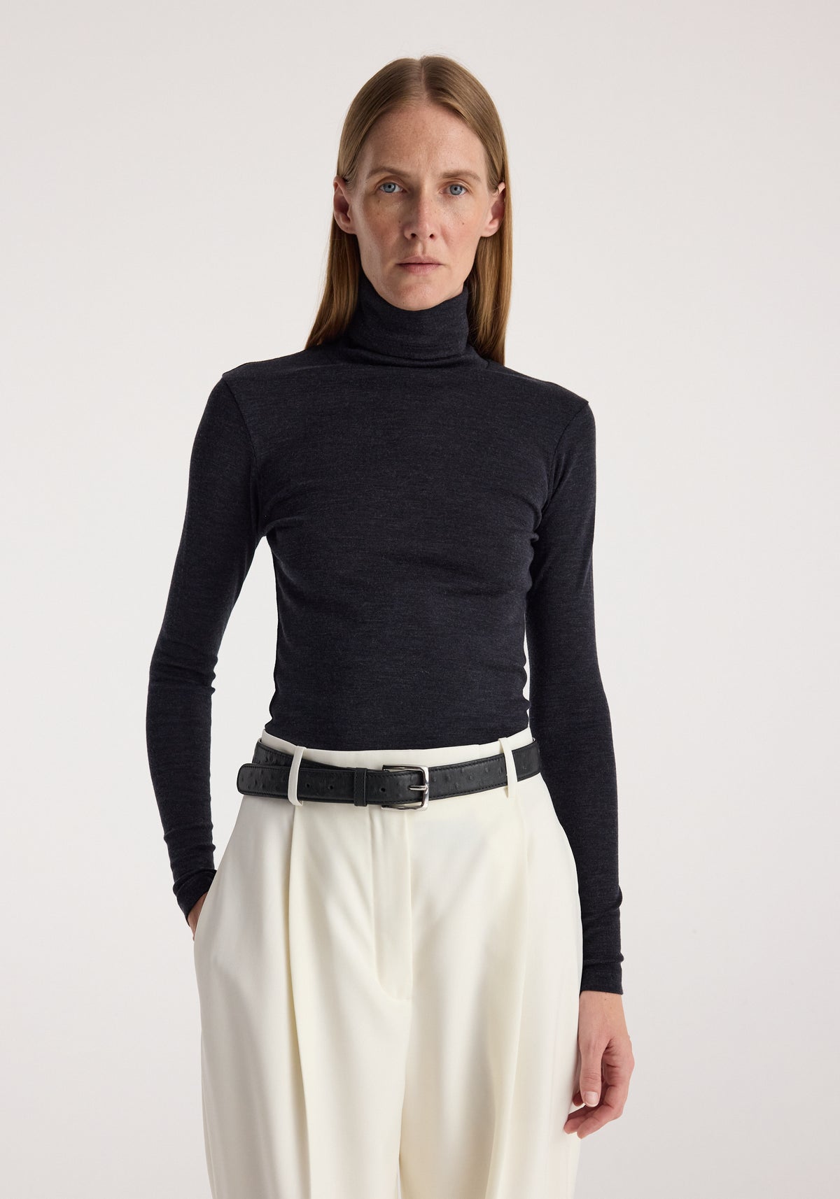 Woman wearing a fitted fine merino turtleneck | anthracite melange with long sleeves, paired with high-waisted white pants and a black belt, standing against a plain background.