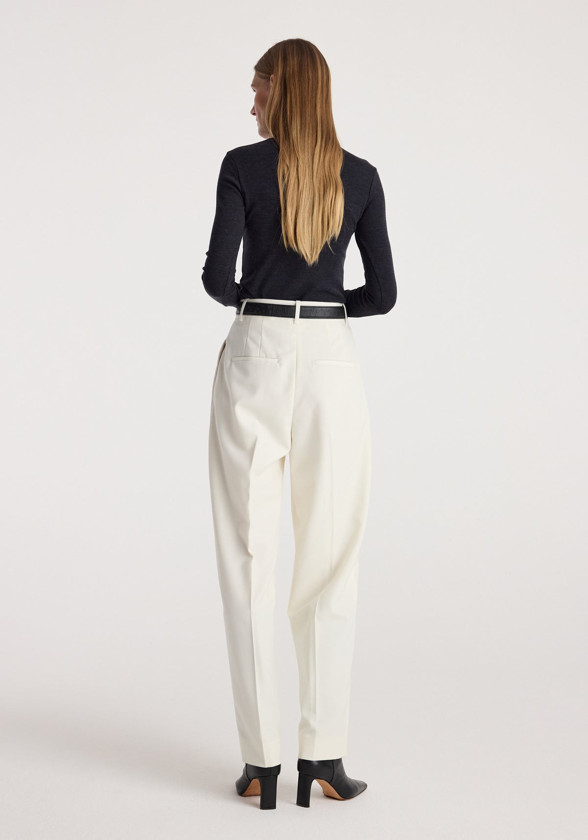 Back view of a woman wearing a fine merino turtleneck | anthracite melange, paired with white high-waisted trousers and black heeled ankle boots against a plain background.