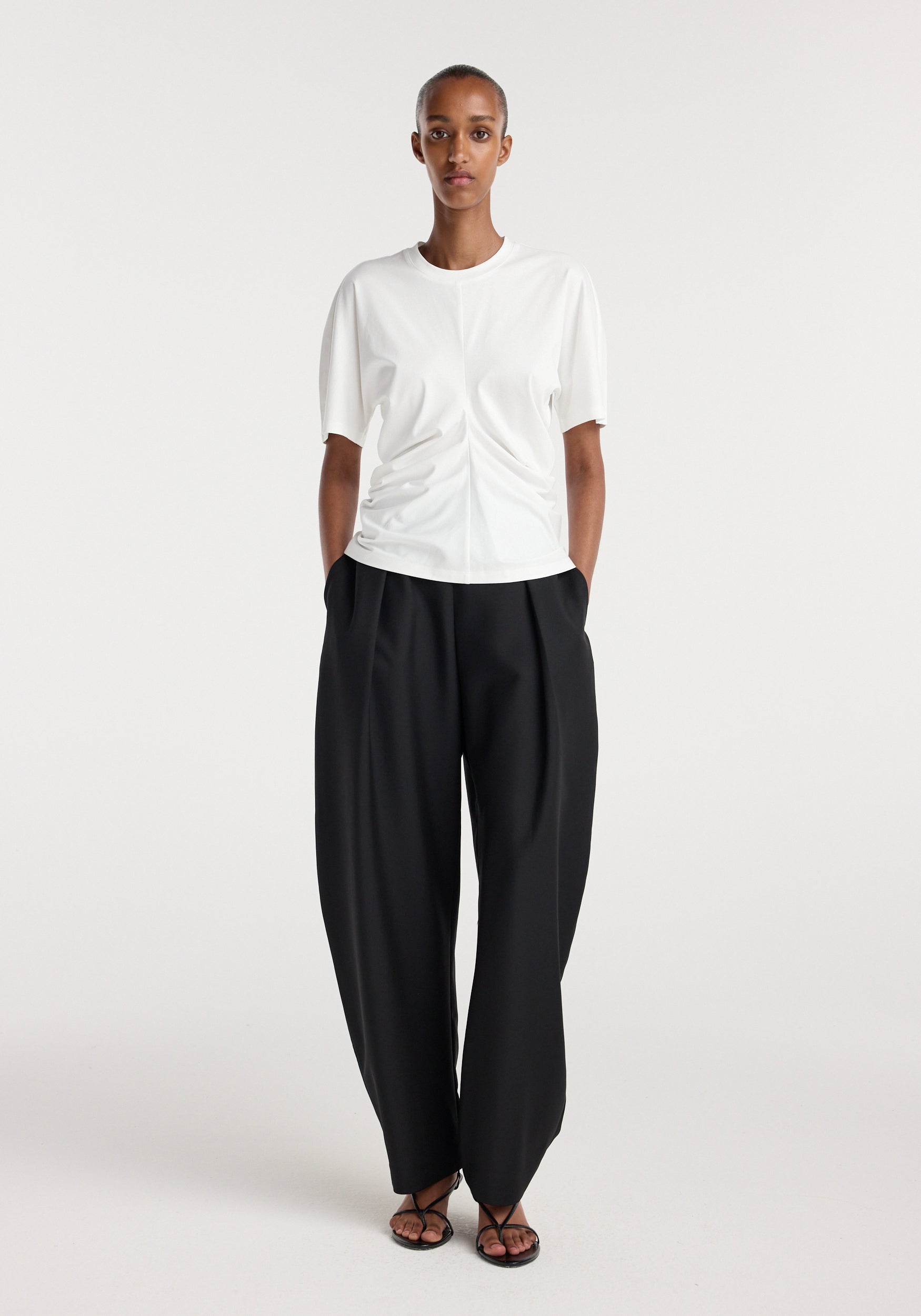 Front view of a model wearing a Draped t-shirt | off white with a gathered detail at the waist, paired with loose black trousers and open-toe sandals, standing against a plain white background.