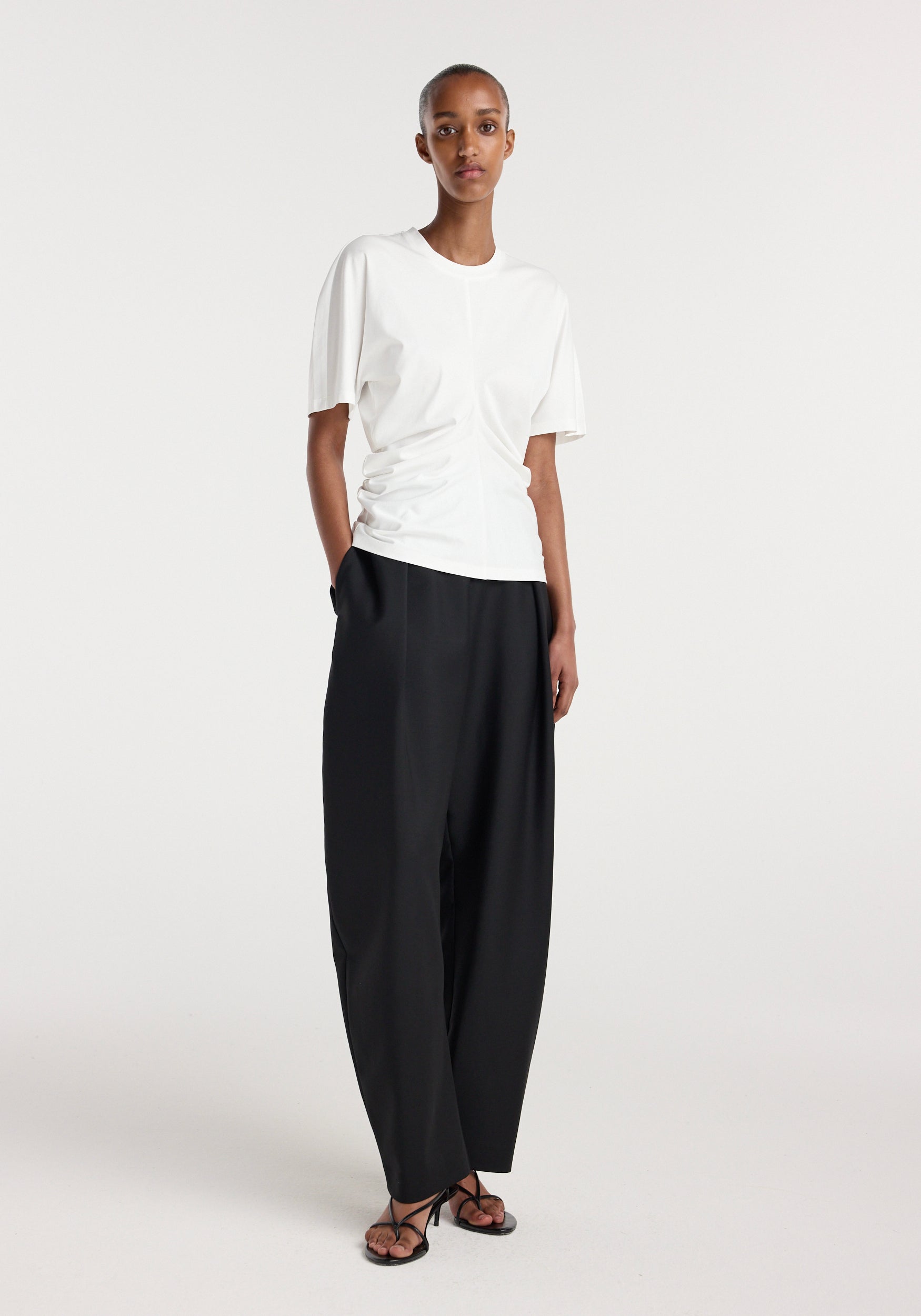 Woman wearing an off white Draped t-shirt | off white with short sleeves and subtle front draping, paired with loose black pants and minimal strappy sandals, standing against a plain background.