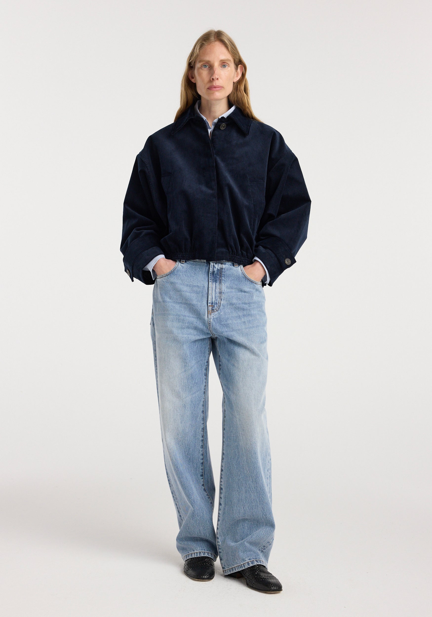 A woman wears a navy blue corduroy blouson jacket | night with hands in pockets, paired with light blue wide-leg jeans and black shoes, standing against a plain background.