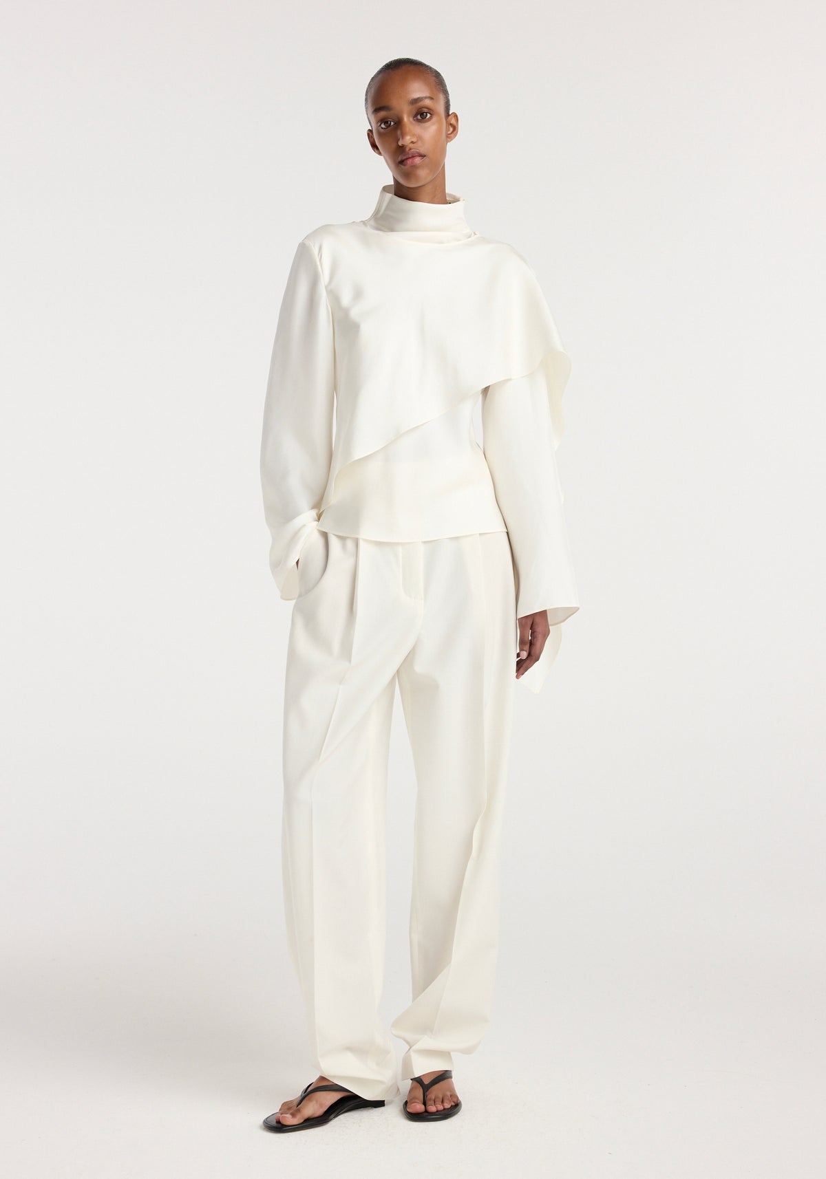 Model wearing a cream-colored Silk foulard top | cream with long sleeves and an asymmetrical draped design paired with matching loose trousers and black sandals.