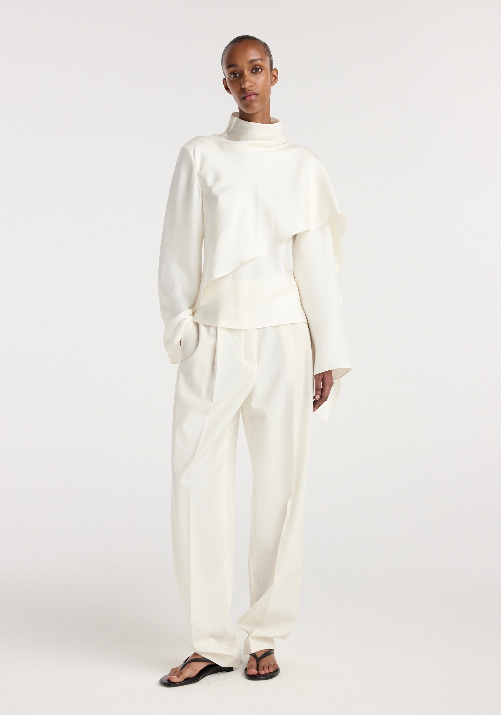 Model wearing a cream-colored Silk foulard top | cream with long sleeves and an asymmetrical draped design paired with matching loose trousers and black sandals.